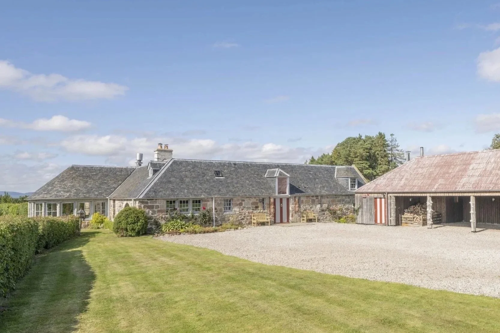 A rustic stone house with a gray shingled roof and multiple chimneys, surrounded by a well-maintained lawn and garden, with a gravel driveway and a wooden carport containing stacked firewood.