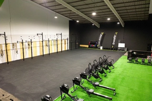 Empty indoor gym with rowing machines, battle ropes, Black background, bright lighting, and green turf for exercises.