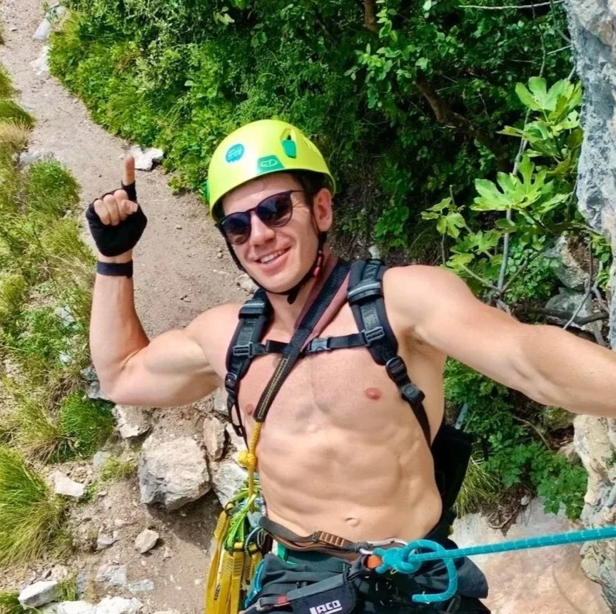 A shirtless man wearing sunglasses, a yellow climbing helmet, and a harness, taking a selfie while rock climbing outdoors amidst greenery.