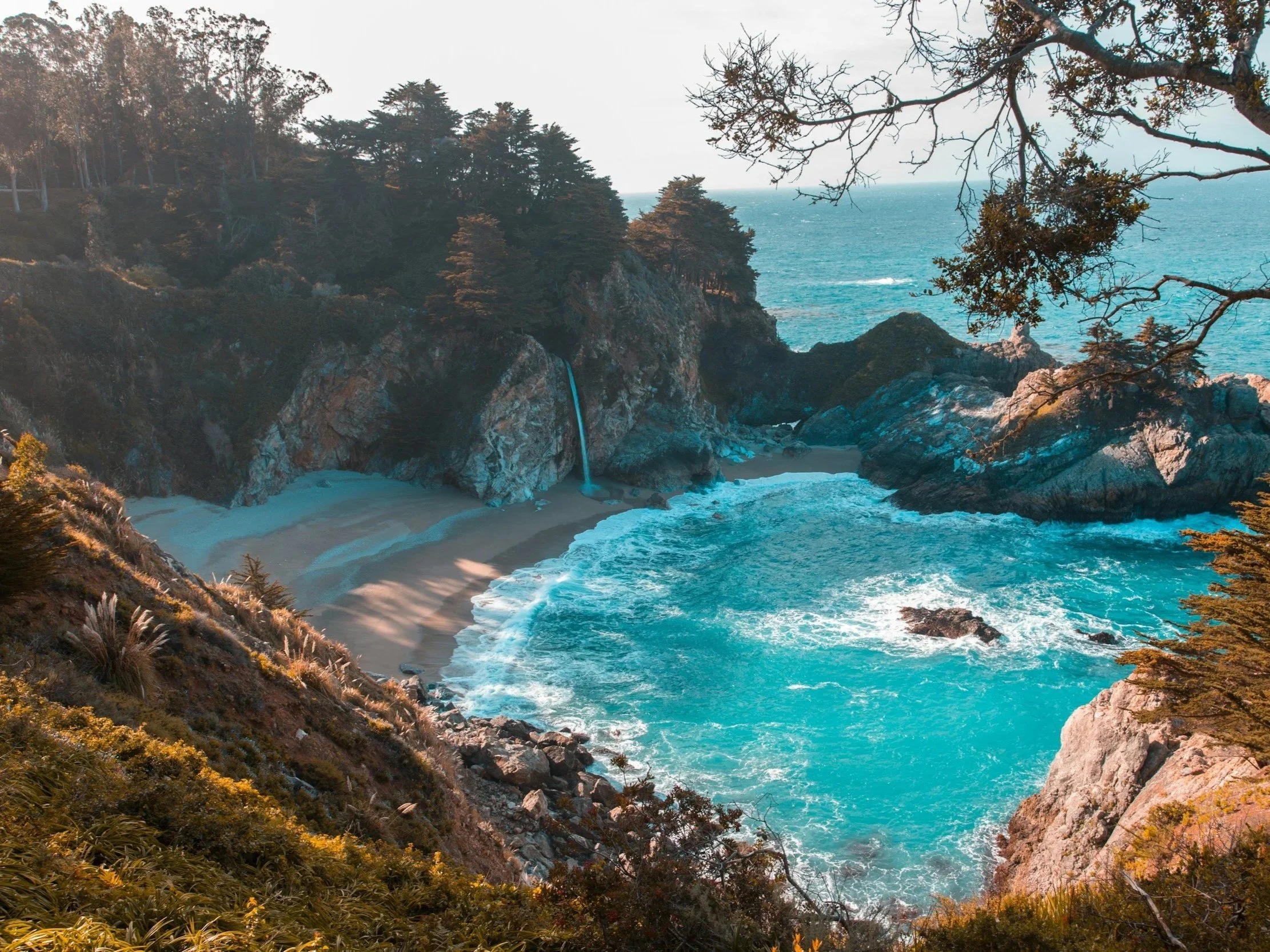 Scenic view of a coastal cove with a waterfall cascading down rock cliffs onto a sandy beach, surrounded by trees and ocean waves.