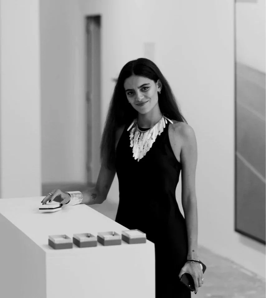 A woman with long dark hair in a sleeveless black dress with a statement necklace, standing at an art gallery with her hand on a small white pedestal displaying four small rectangular objects.