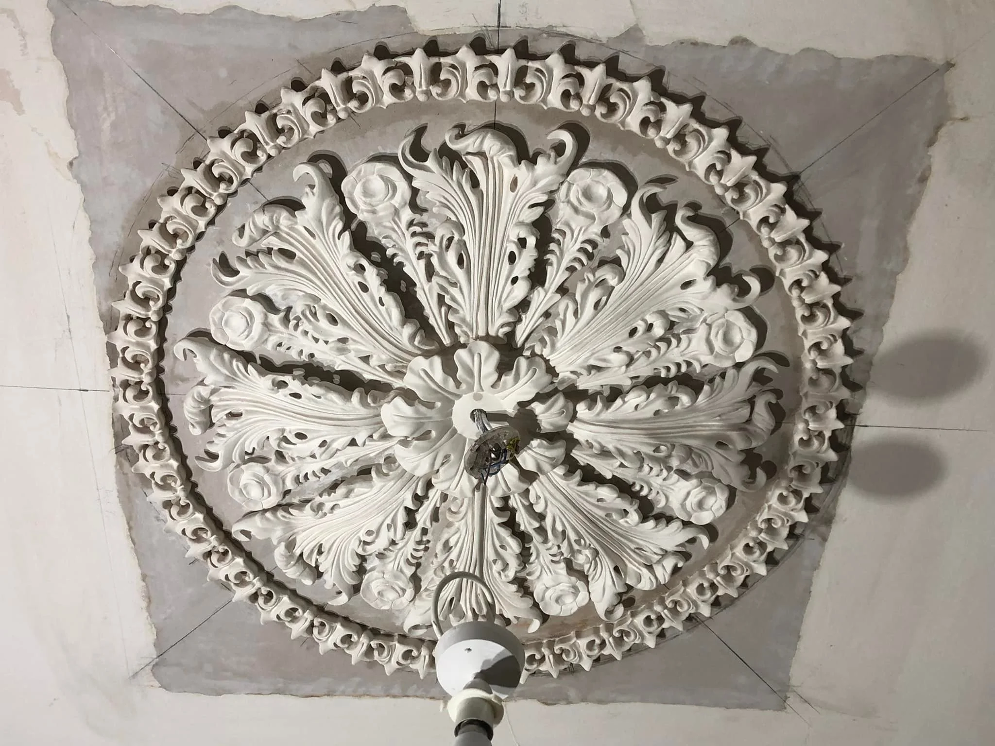 Ornate white ceiling medallion with floral and scroll patterns, partially installed around a hanging light fixture in an unfinished room.