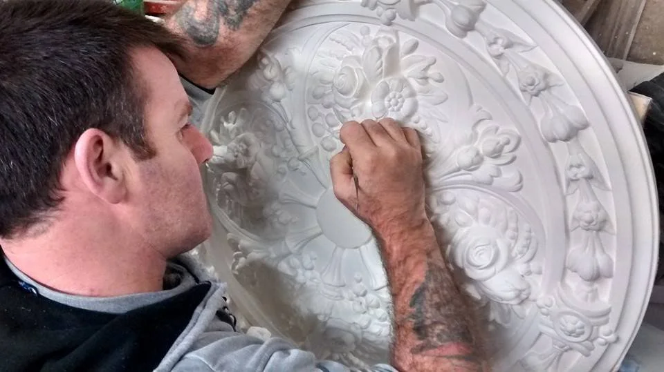Man carving intricate floral design into a white decorative ceiling medallion.