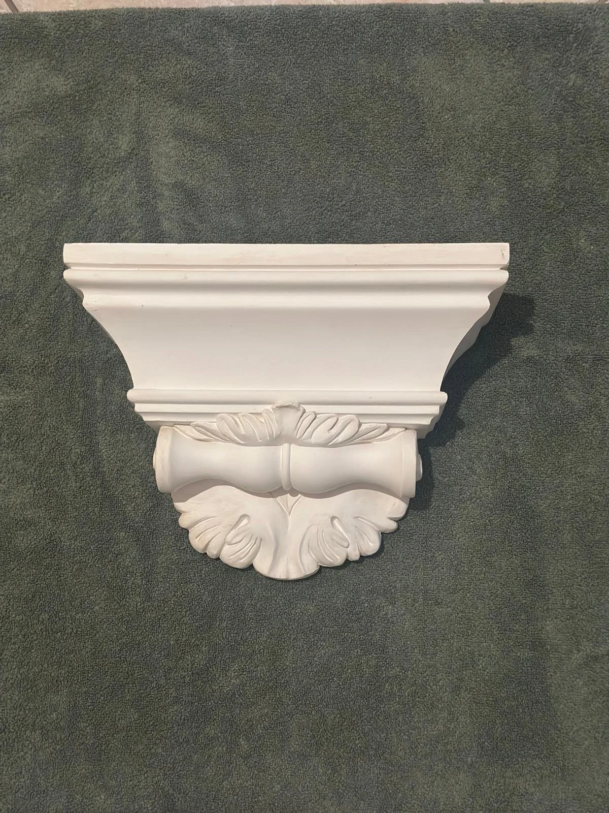 A decorative white wall corbel with ornate leaf and scroll design against a dark green textured background.