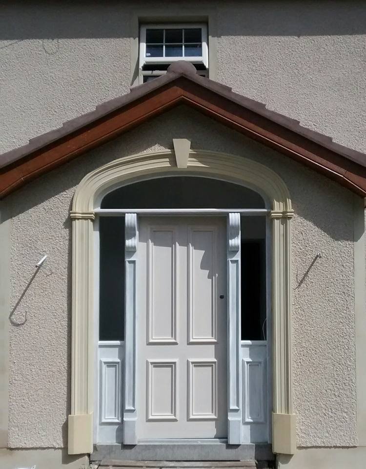 Front entrance of a house with a white door, decorative trim, and an arched window above, under a gabled roof.