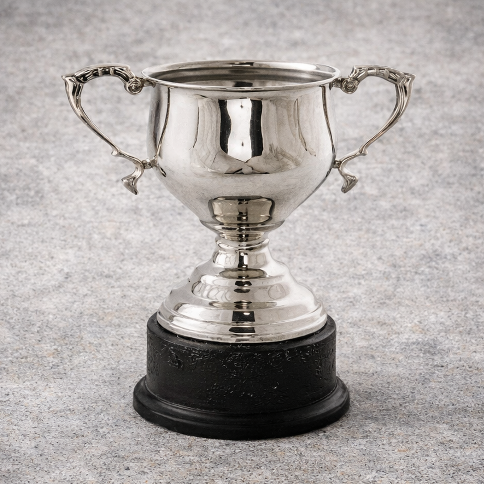 Silver trophy cup with ornate handles on a black base, placed on a gray, textured surface.