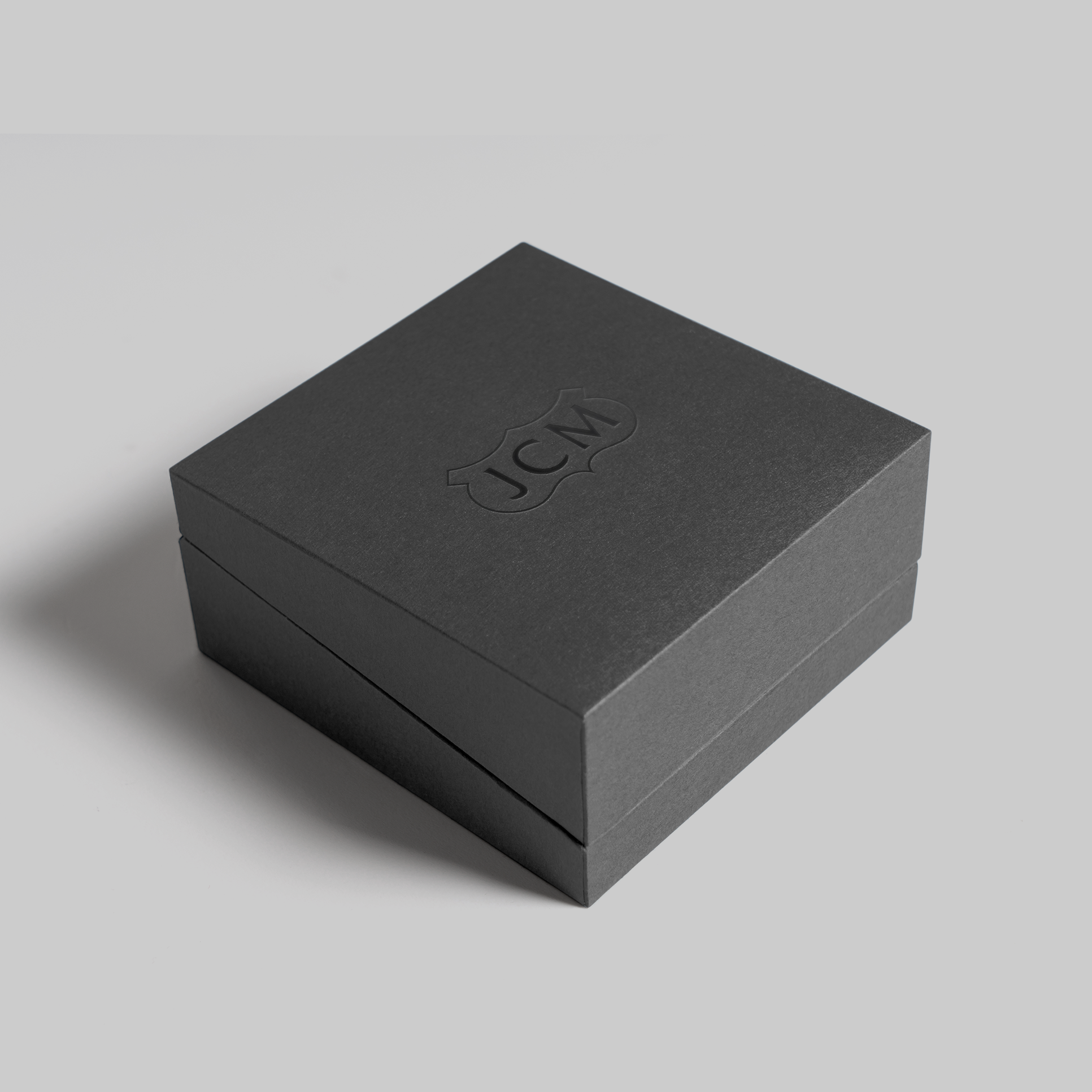 A black jewelry box with embossed logo 'JCM' on the lid, placed on a light gray surface.