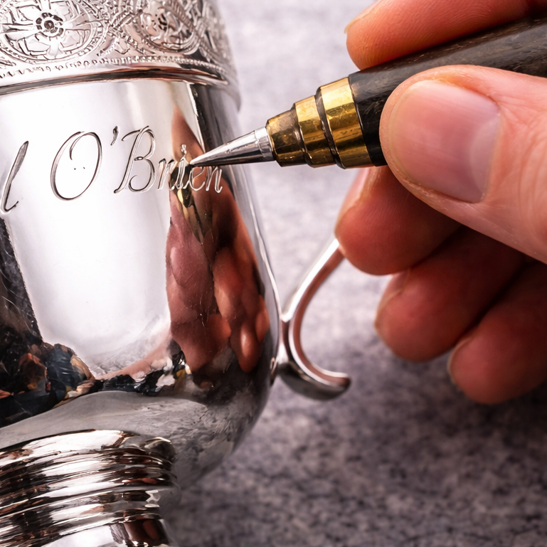 A person is engraving the name "L O'Brien" on a shiny silver trophy with a metal engraving pen.