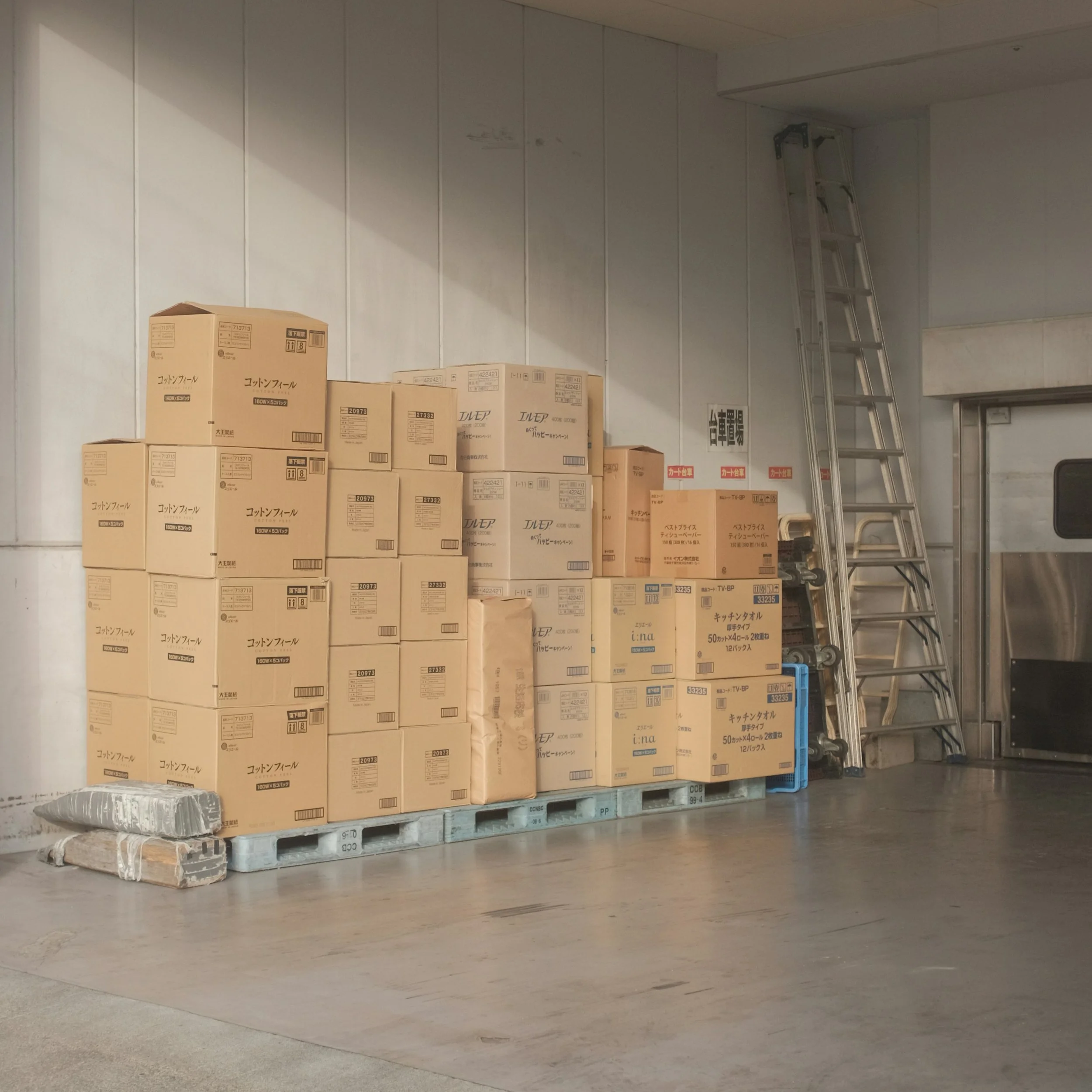 A stack of cardboard boxes on a wooden pallet next to a metal ladder in a warehouse. The boxes have Japanese writing and are in front of a white wall.
