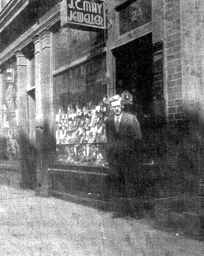 A man in a suit standing outside a jewelry store called J.C. May Jeweller, with a display of jewelry and other items in the window, on a city street.