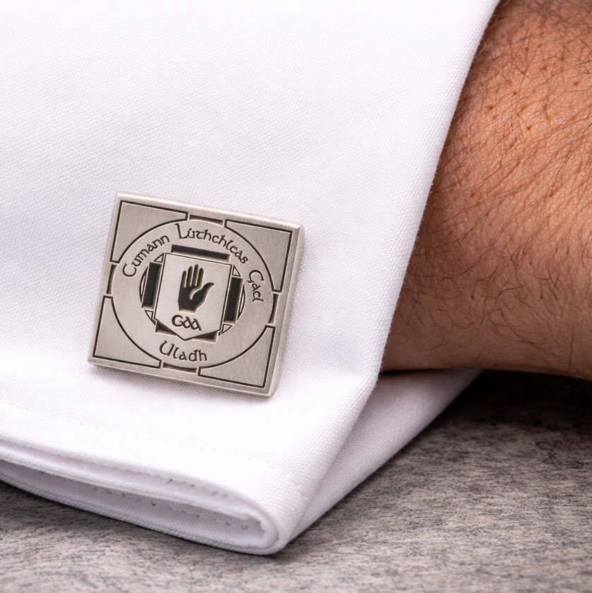 A close-up of a cufflink on a white shirt sleeve. The cufflink has an Irish design with the words 'Cumann Lúthchleas Gael' and a black hand symbol in the center.