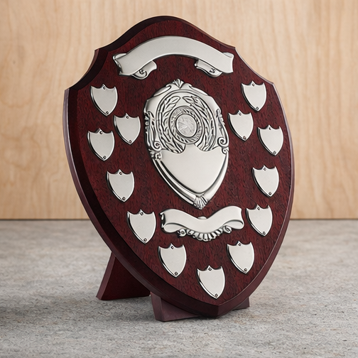 A wooden shield-shaped trophy plaque with multiple small shield-shaped metal plates around a central larger plate, all sitting on a flat surface against a wooden background.