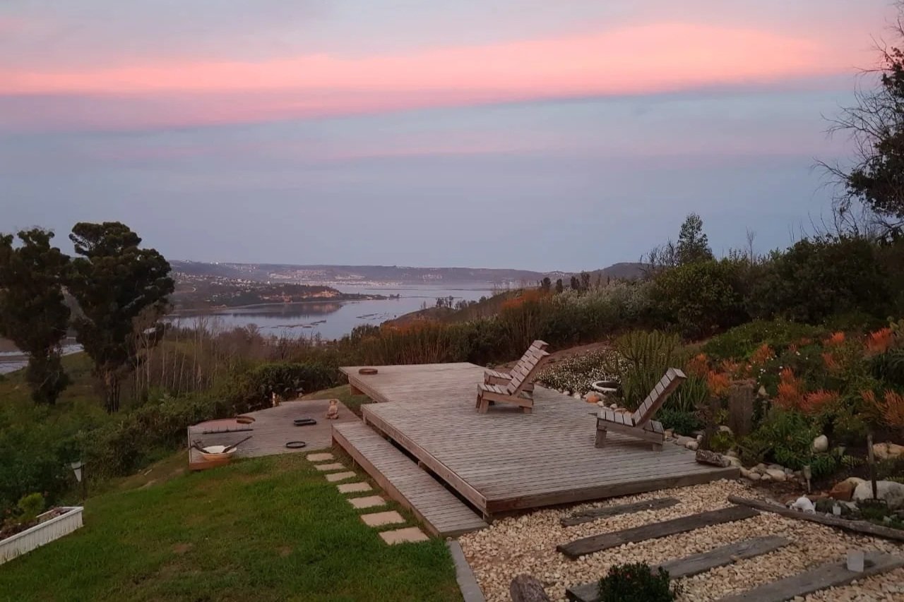 A backyard deck with Adirondack chairs overlooking a landscape of trees and a river at sunset, with a colorful sky, designed and installed by Outdoor Spaces team.