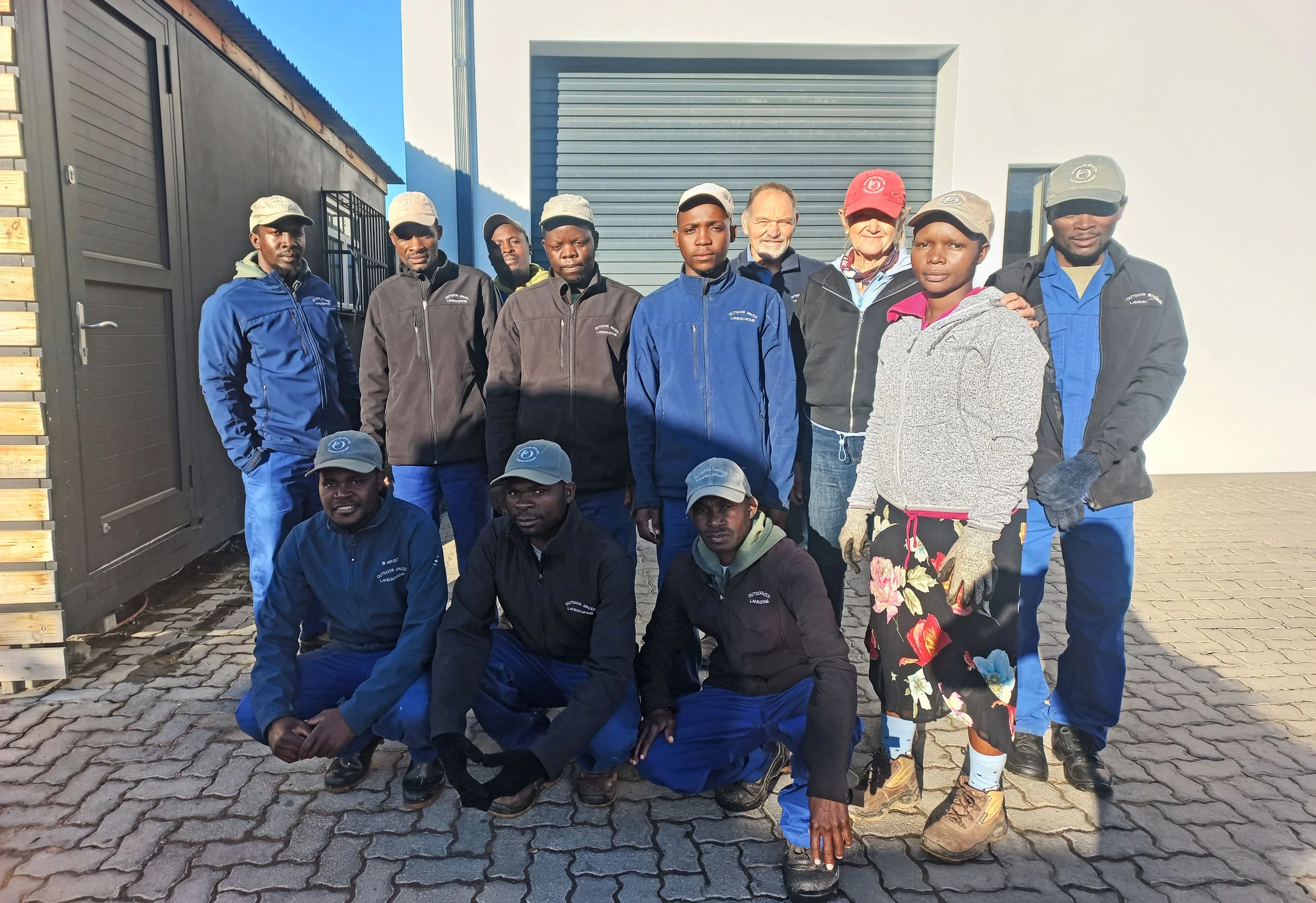 Outdoor Spaces Team, posing for a photo in front of a building with a gray metal door. Some are standing, some are crouching, and one woman is with her arm around a man. They are dressed in outdoor clothing, ready for work.
