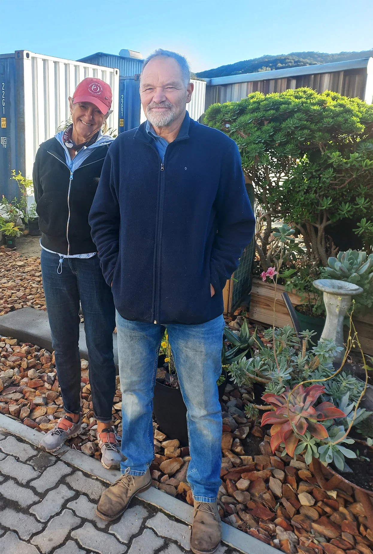 Outdoor Spaces co founders, Margi and Jeremy. Both smiling, standing outdoors in a garden with plants, rocks, and container planters, with a blue sky and mountains in the background.