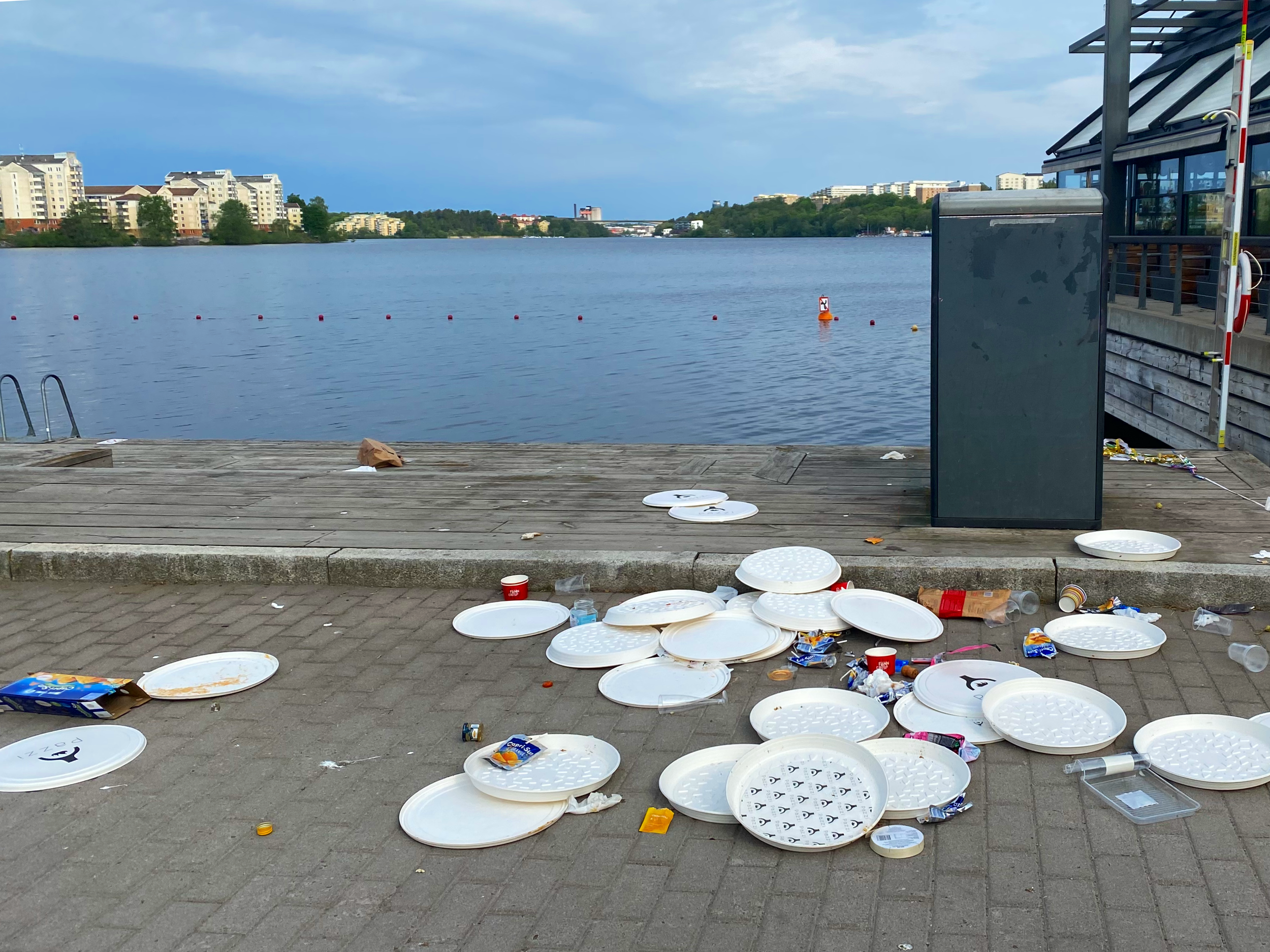 Flertal engångspappersfat och annat förpackningsskräp på en kaj nere vid vattnet, nära en soptunna.