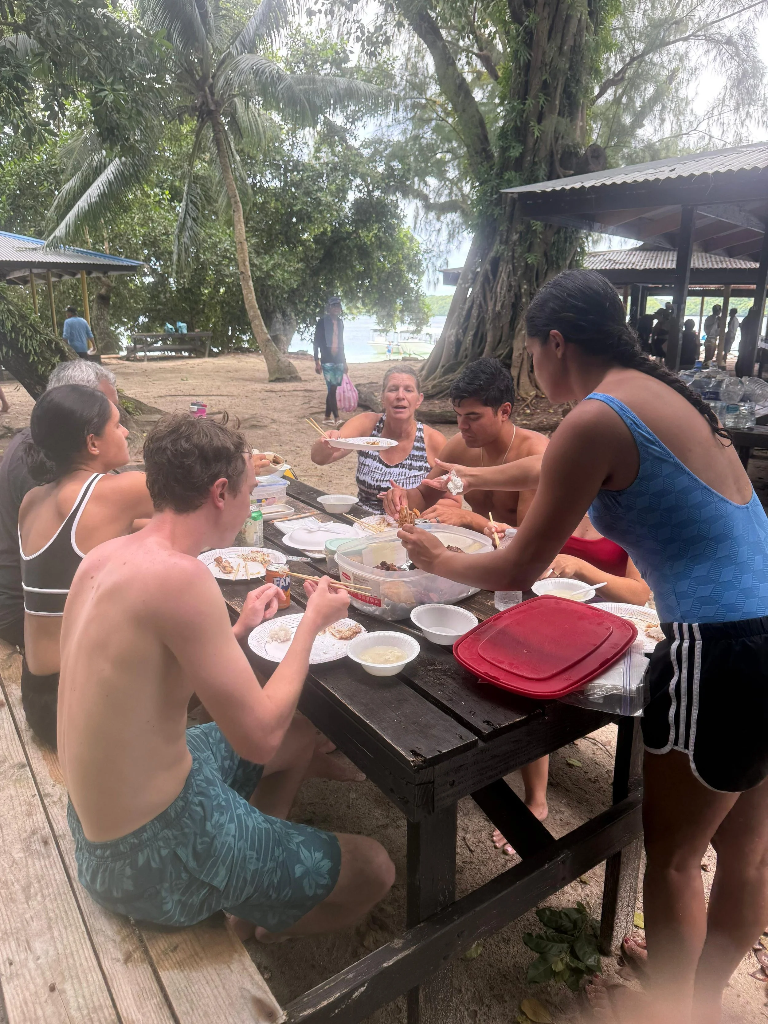 People gathered around a table outdoors, enjoying a meal near a beach with large trees and a thatched shelter in the background.