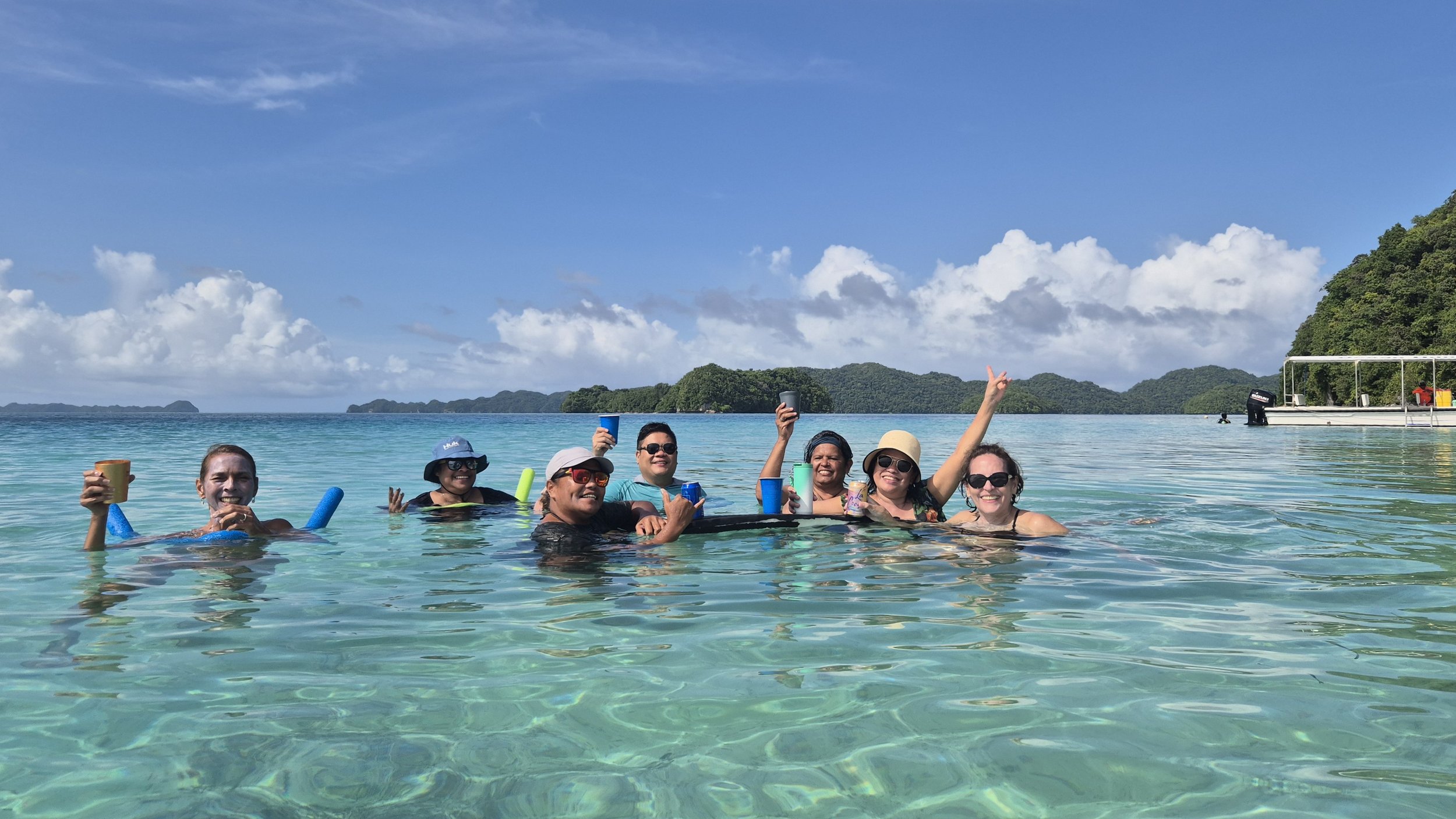 Group of eight people swimming and relaxing in clear blue water near a tropical island, holding drinks and enjoying sunny weather.