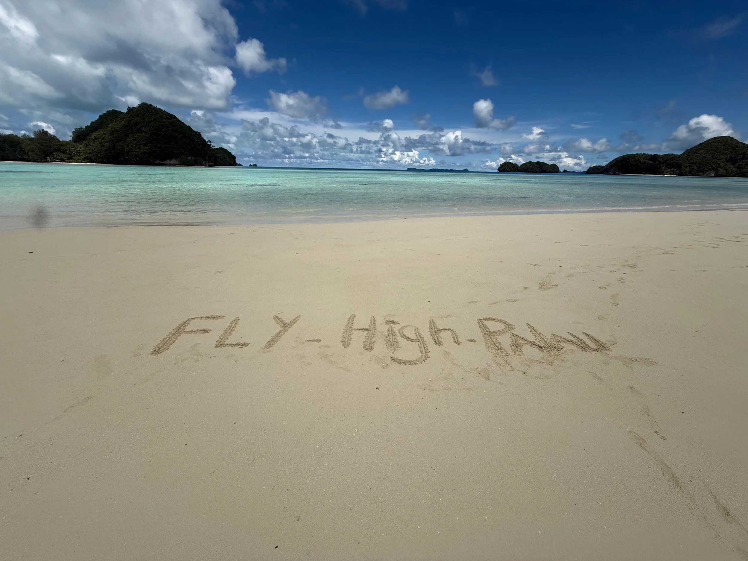 Sand on the beach with the words 'FLY-High-PNW' written in the sand, overlooking clear turquoise water and green islands under a partly cloudy sky.