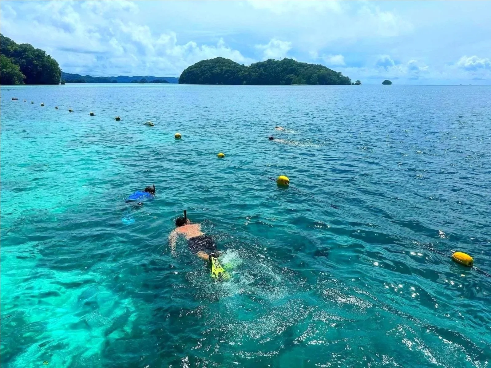 Two snorkelers swimming in clear blue ocean water near a chain of yellow and white buoys, with green islands and a partly cloudy sky in the background.