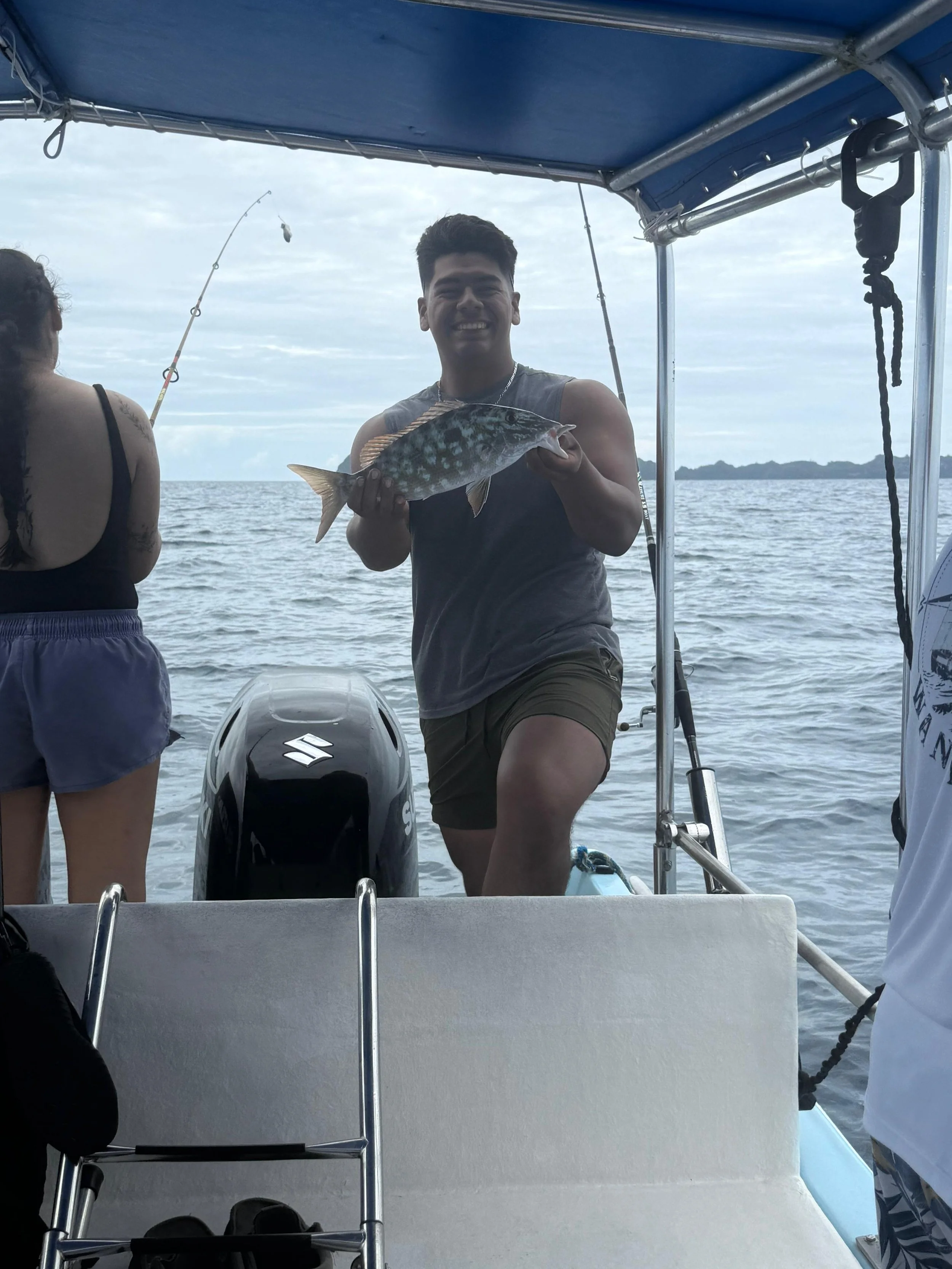 A young man holding a fish on a boat with the ocean and distant islands in the background.