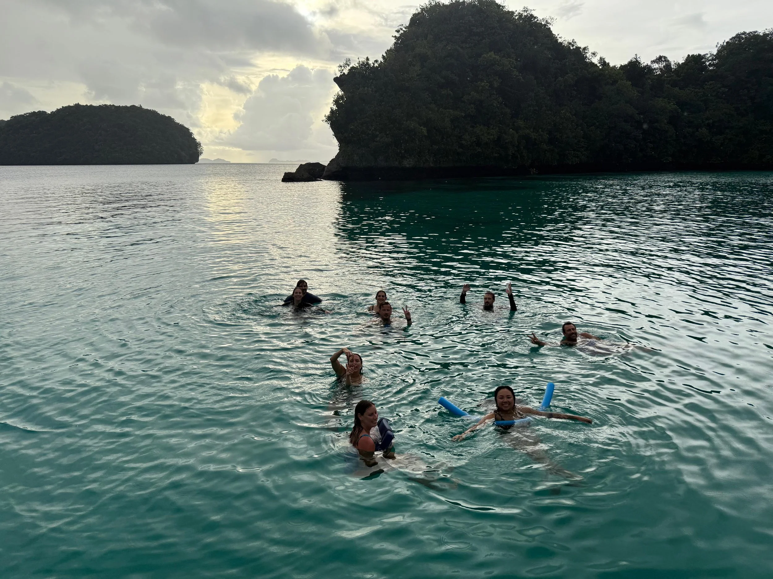 Group of people swimming and relaxing in a tropical bay with lush islands and cloudy sky in the background.