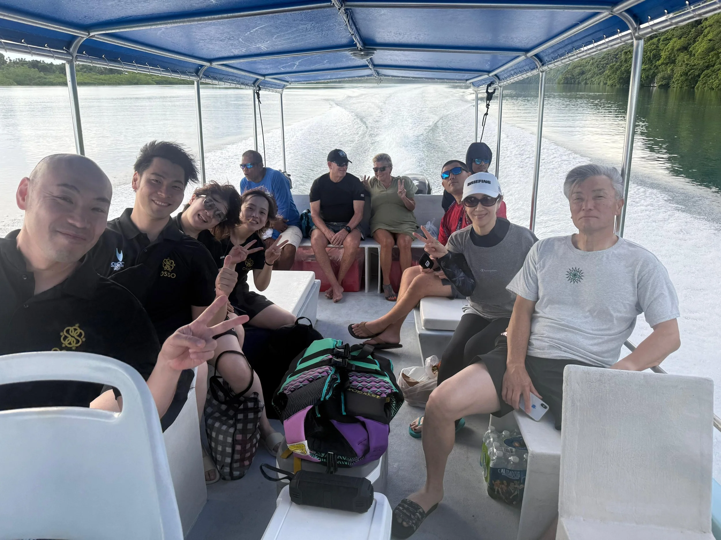 A group of people on a boat with a wake behind them, surrounded by water and green trees, enjoying a sunny day.