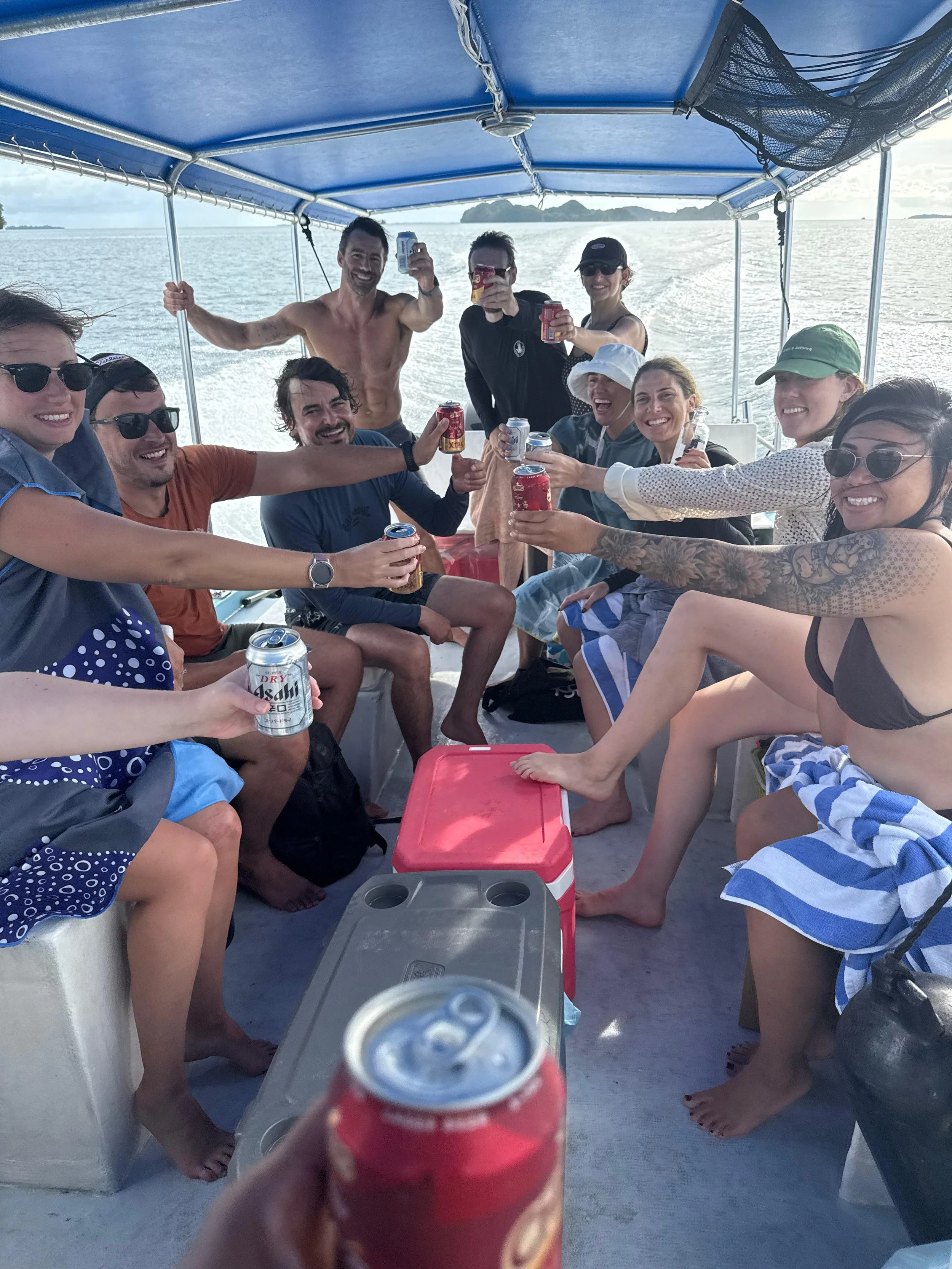 A group of friends on a boat, toasting with canned drinks, smiling, and enjoying a day on the water with a scenic ocean background.