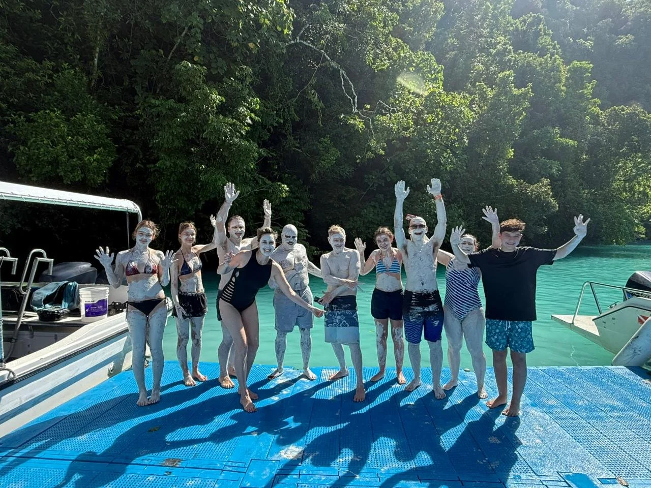 Group of people at a lake, covered in white mud, standing on a blue dock with trees in the background, smiling and waving.