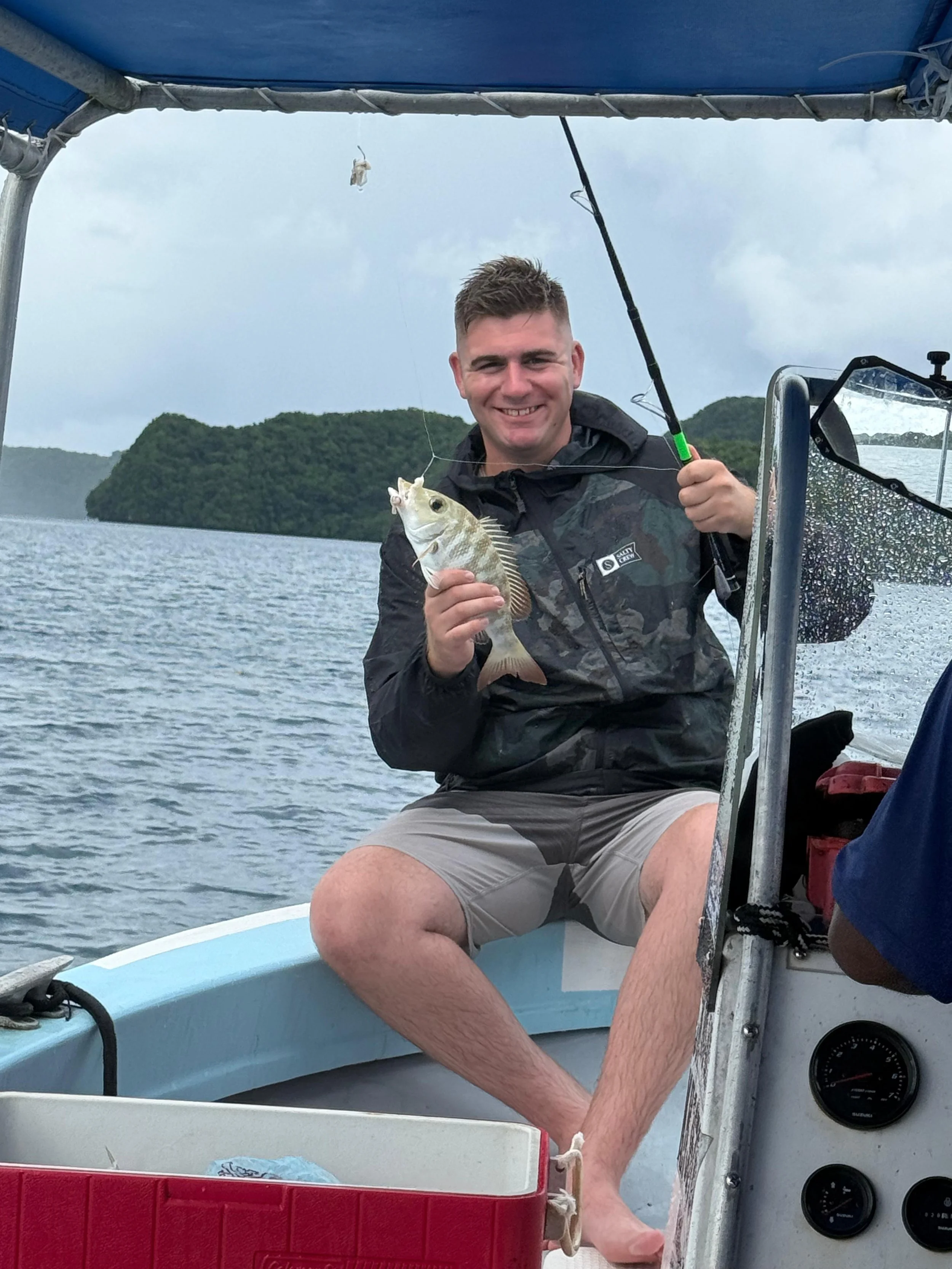 Young man smiling and sitting on a boat holding a small fish he caught, with water and green hills in the background.
