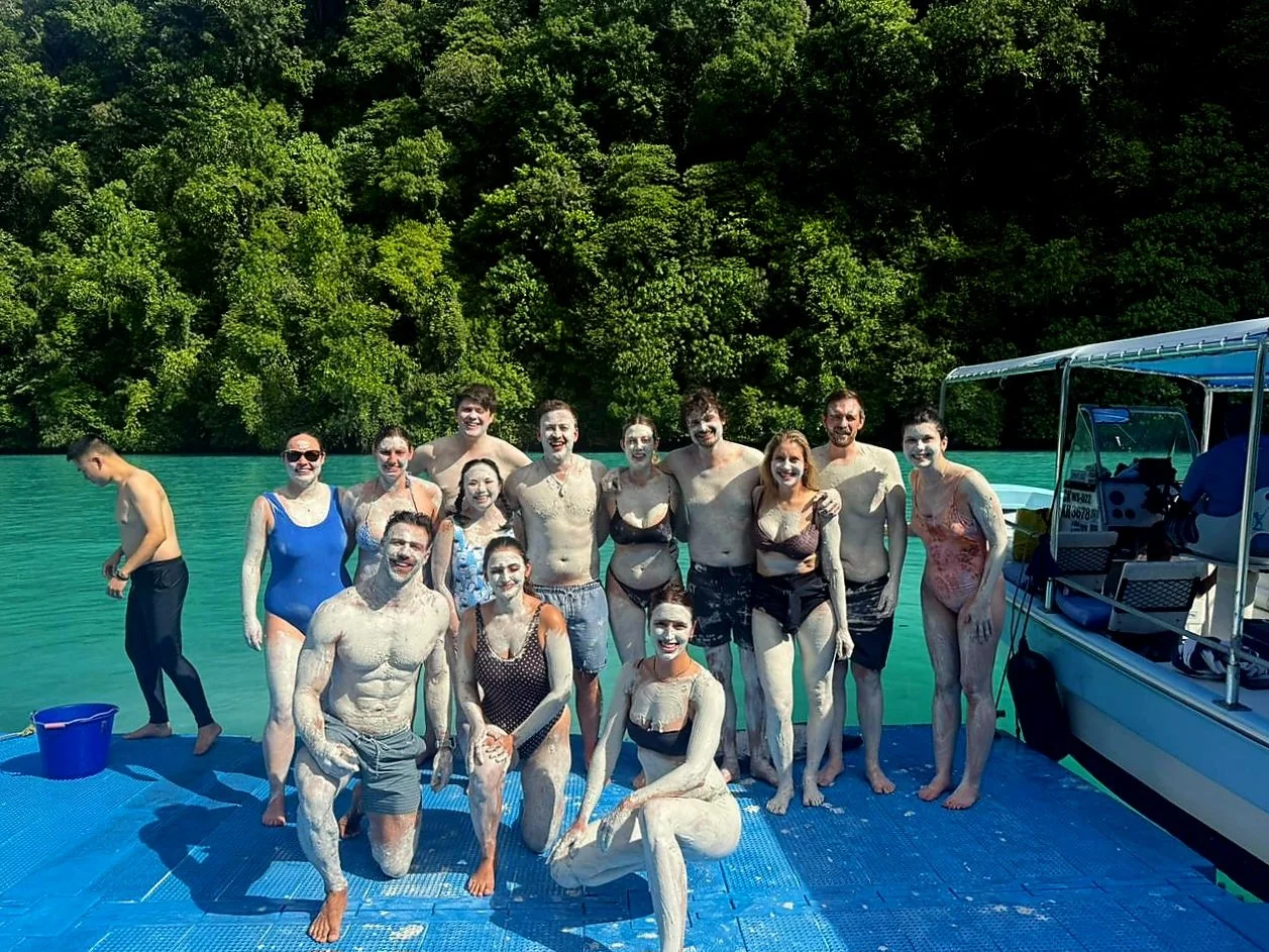 Group of people smiling and covered in foam on a blue floating dock near a boat, with green trees and a lake in the background.