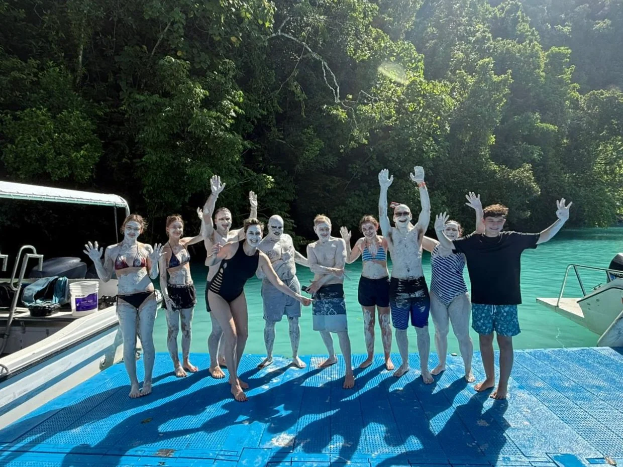 Group of people covered in mud on a dock near water, with green trees in the background, all smiling and raising their hands.