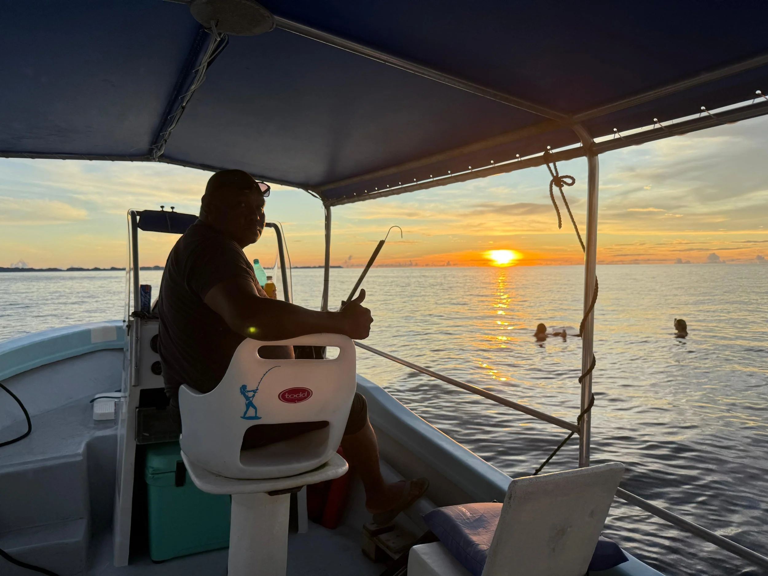 A person sitting on a boat at sunset, holding a fishing rod, with a few people swimming in the water nearby.