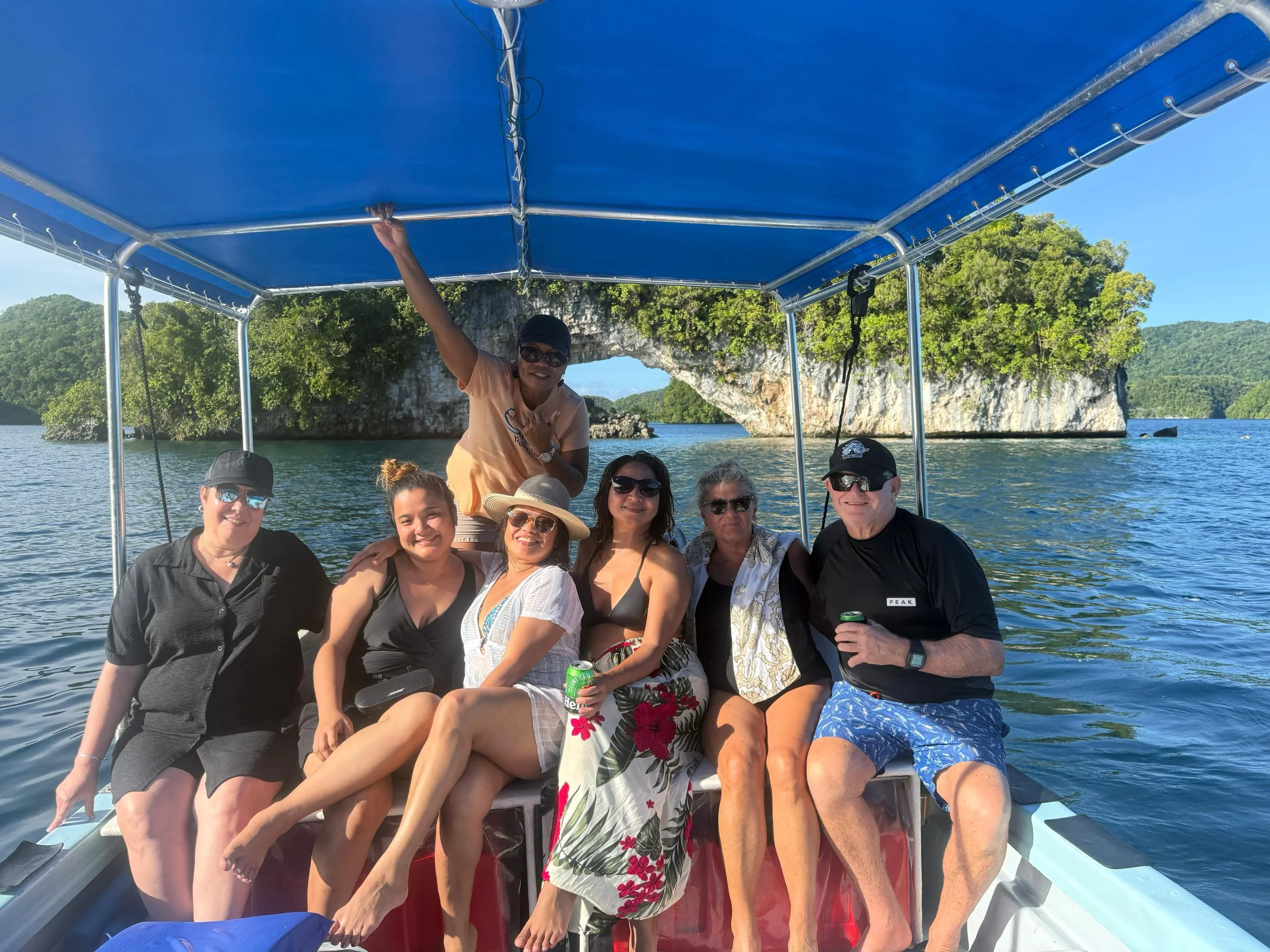 Group of people enjoying a boat ride on a sunny day, with clear water, lush green rocky islands, and an arch-shaped rock formation in the background.