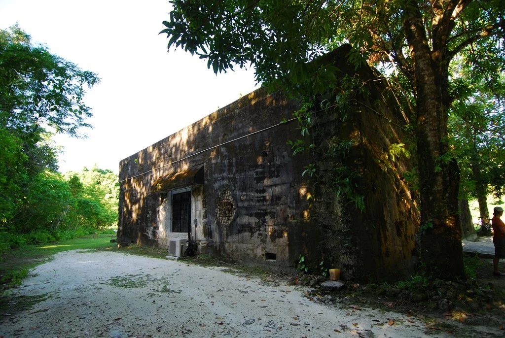 An old, weathered building made of dark stone, surrounded by lush green trees and foliage, with a dirt pathway in front and a person standing nearby.