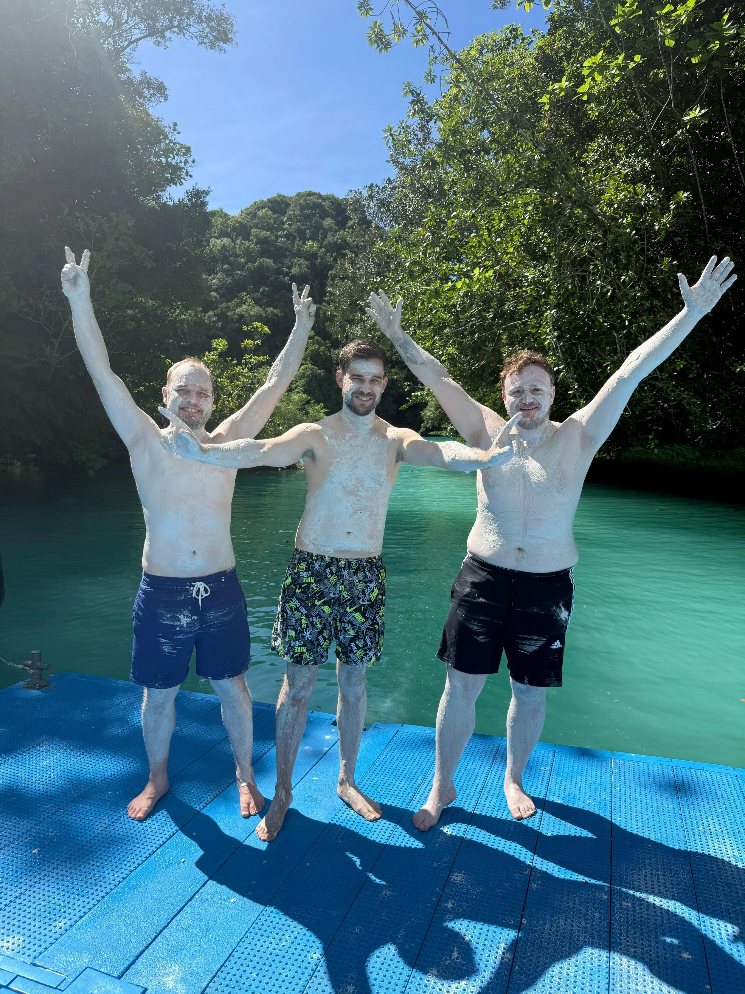Three men smiling with arms raised, covered in white chalk or powder, standing on a blue dock by a body of water surrounded by green trees on a sunny day.