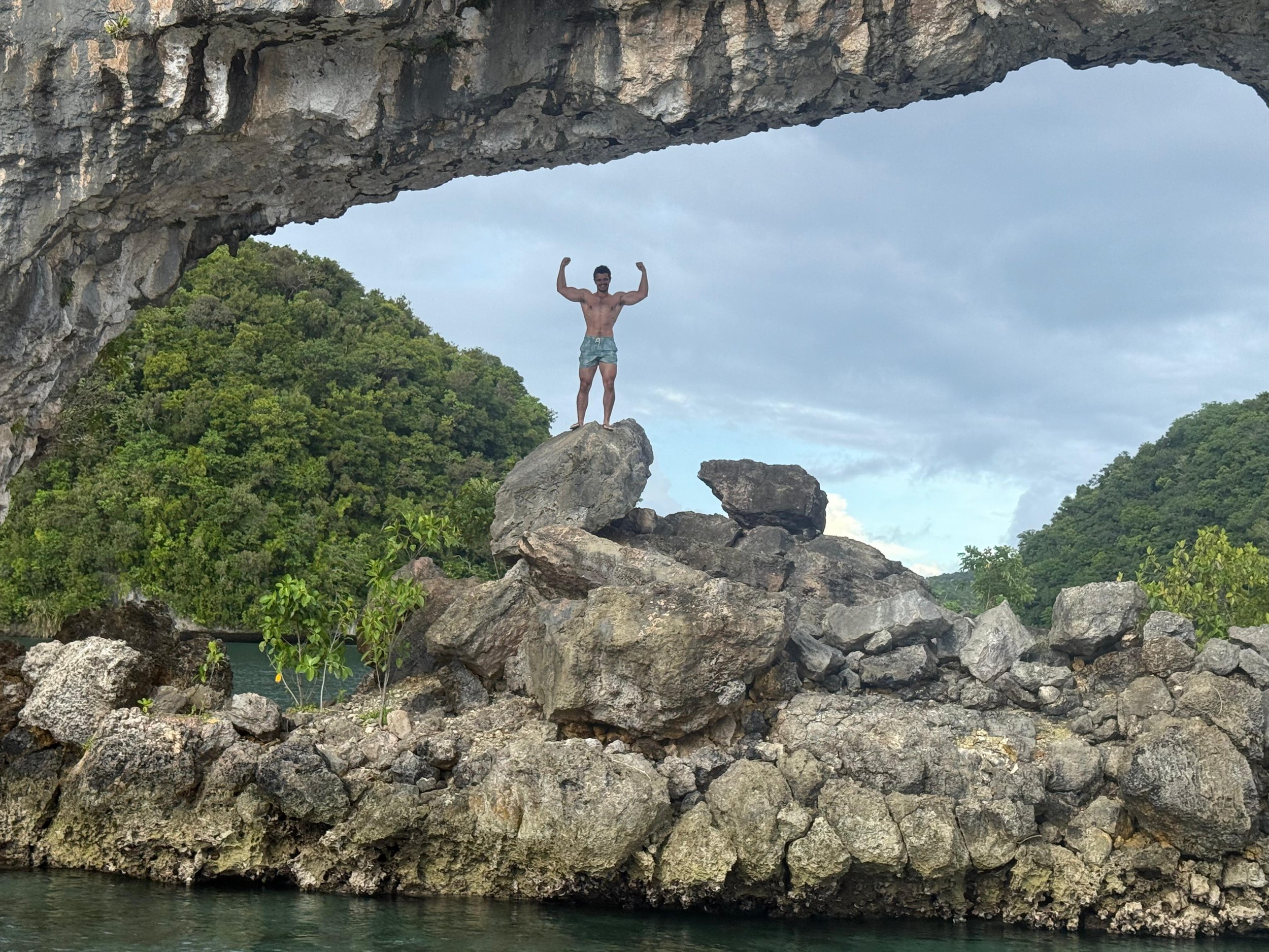 A shirtless man in shorts standing on a large rock formation with his arms raised in a flexing pose, beneath an overhanging rock in a lush, green landscape with trees and hills, near a body of water.
