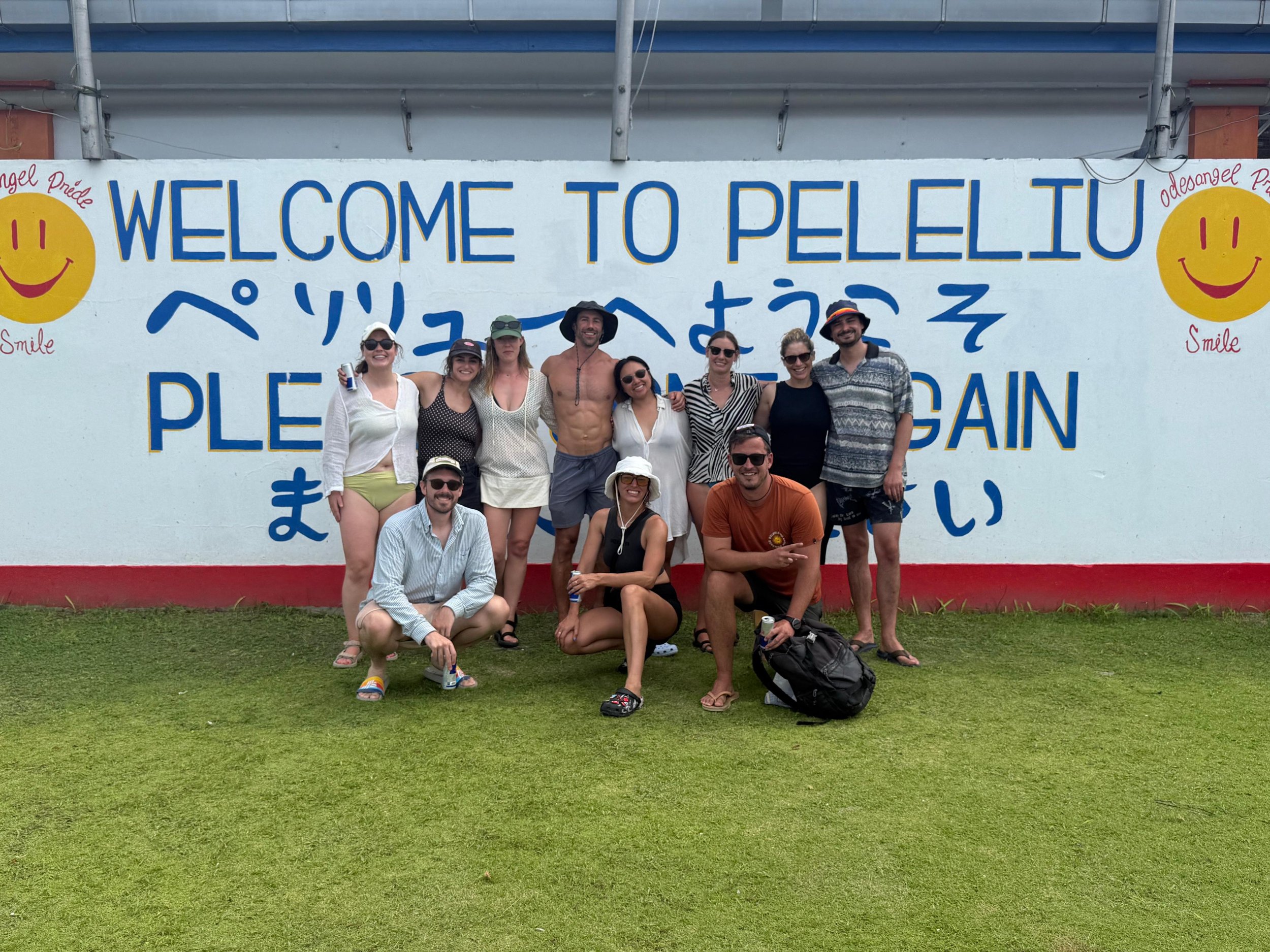 Group of ten people posing in front of a large wall with colorful sign that says "Welcome to Pelelli" in English and Japanese, some smiling, wearing summer clothes and sunglasses, with a grassy ground in front.