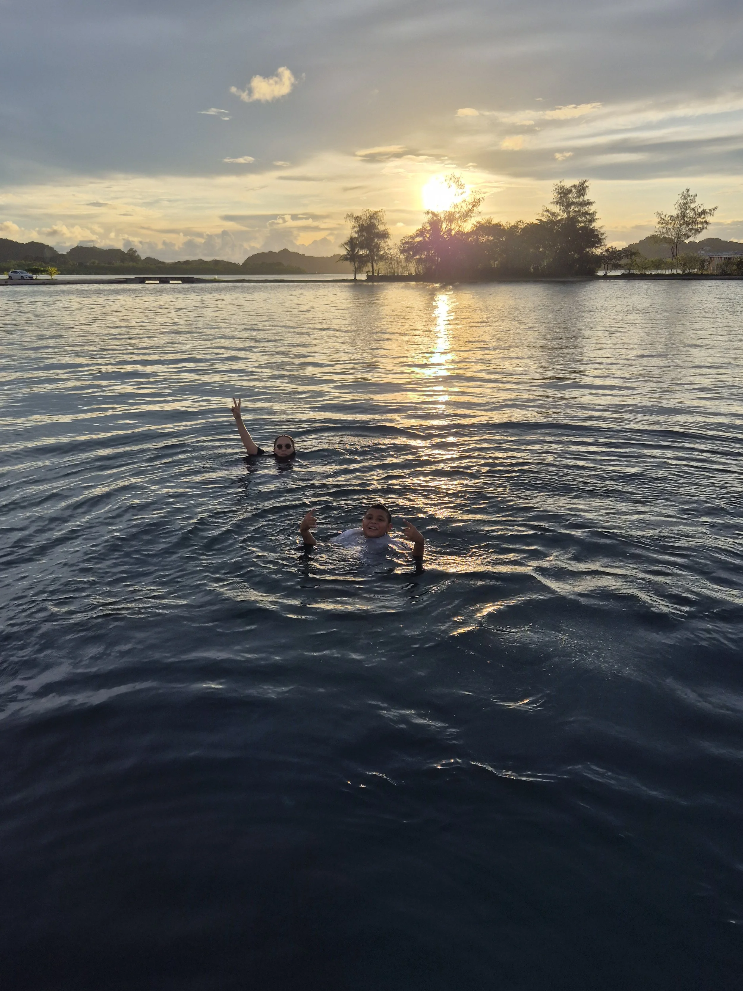 Two children swimming in a lake at sunset, with trees and hills in the background.