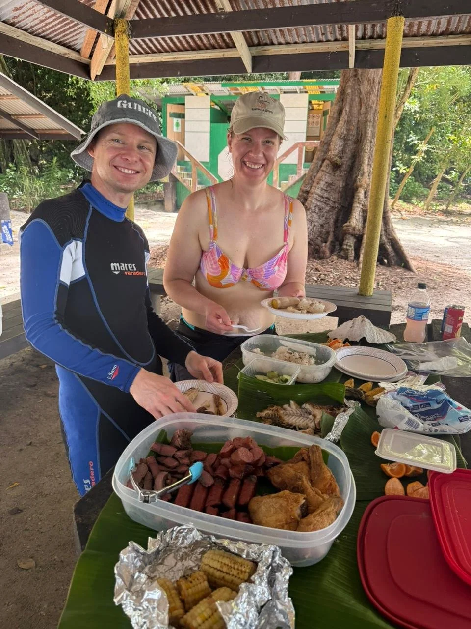 Two people smiling at an outdoor meal under a shelter, with a large tree in the background. The woman is in a bikini top, holding a plate of food, and the man is in a wetsuit with a hat, standing near a table filled with various dishes, including grilled meats, vegetables, and corn. There is a bottle of water and other food containers on the table.