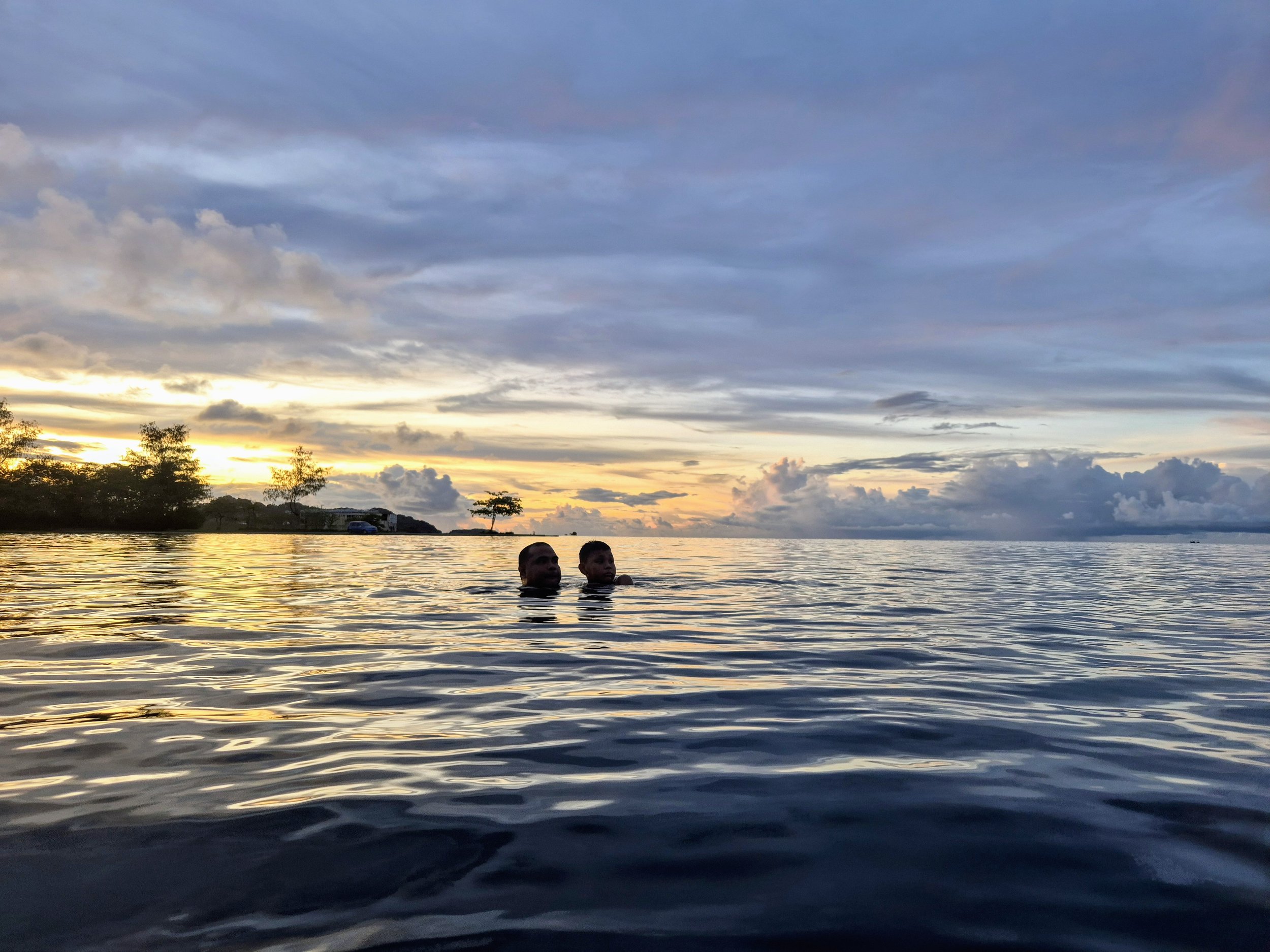 Two people swimming in a large body of water at sunset, with a cloudy sky and some trees on the horizon.