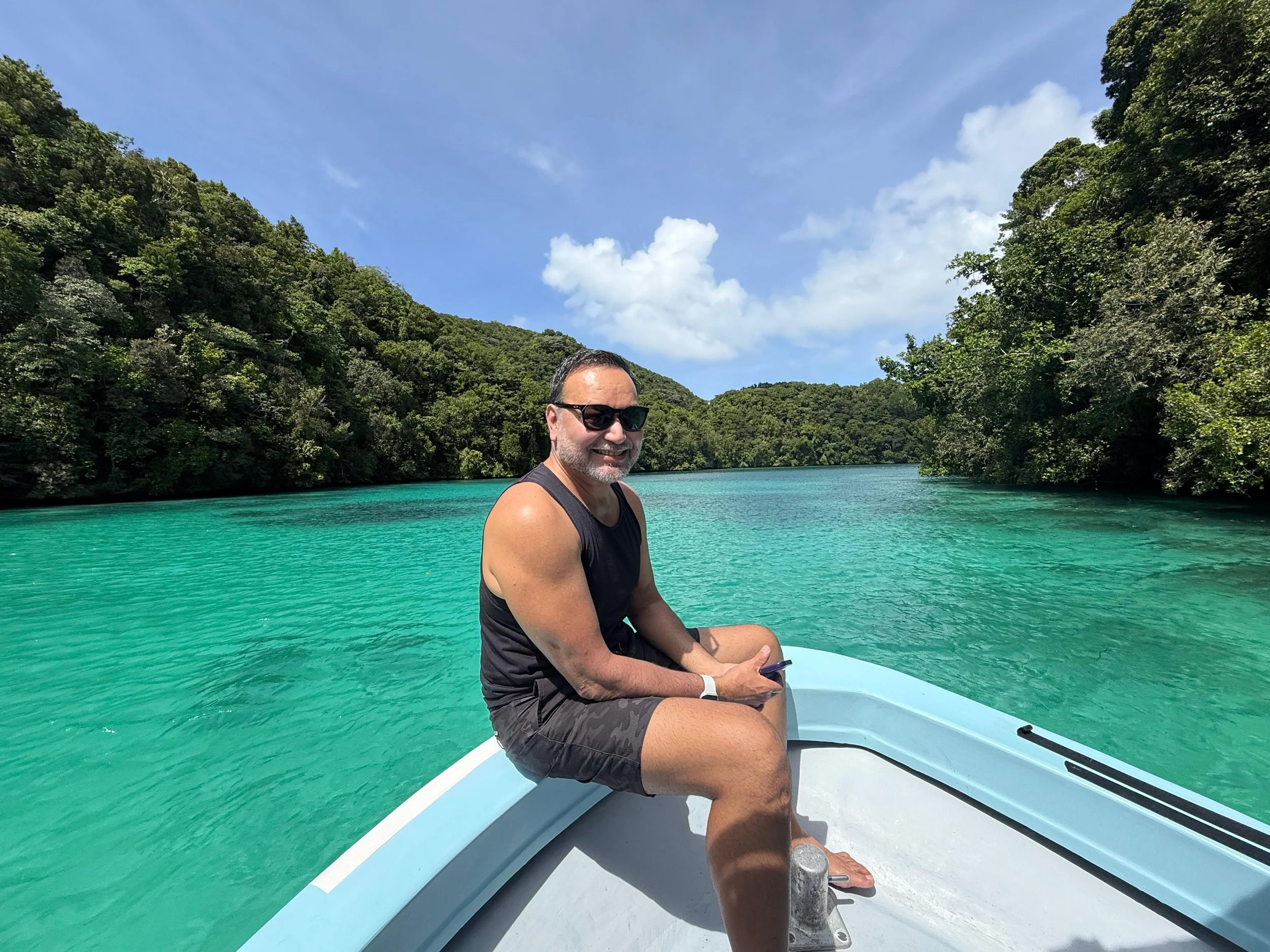 A man with sunglasses and a sleeveless shirt sitting on the edge of a boat, smiling with a view of turquoise water and green forested hills under a partly cloudy sky.