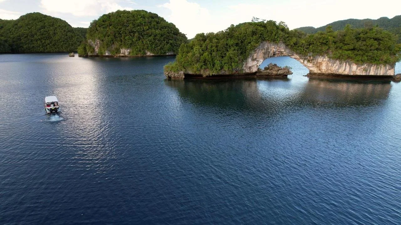 A boat sailing near lush green islands with a natural rock arch and other small islands in the background, on a body of water.