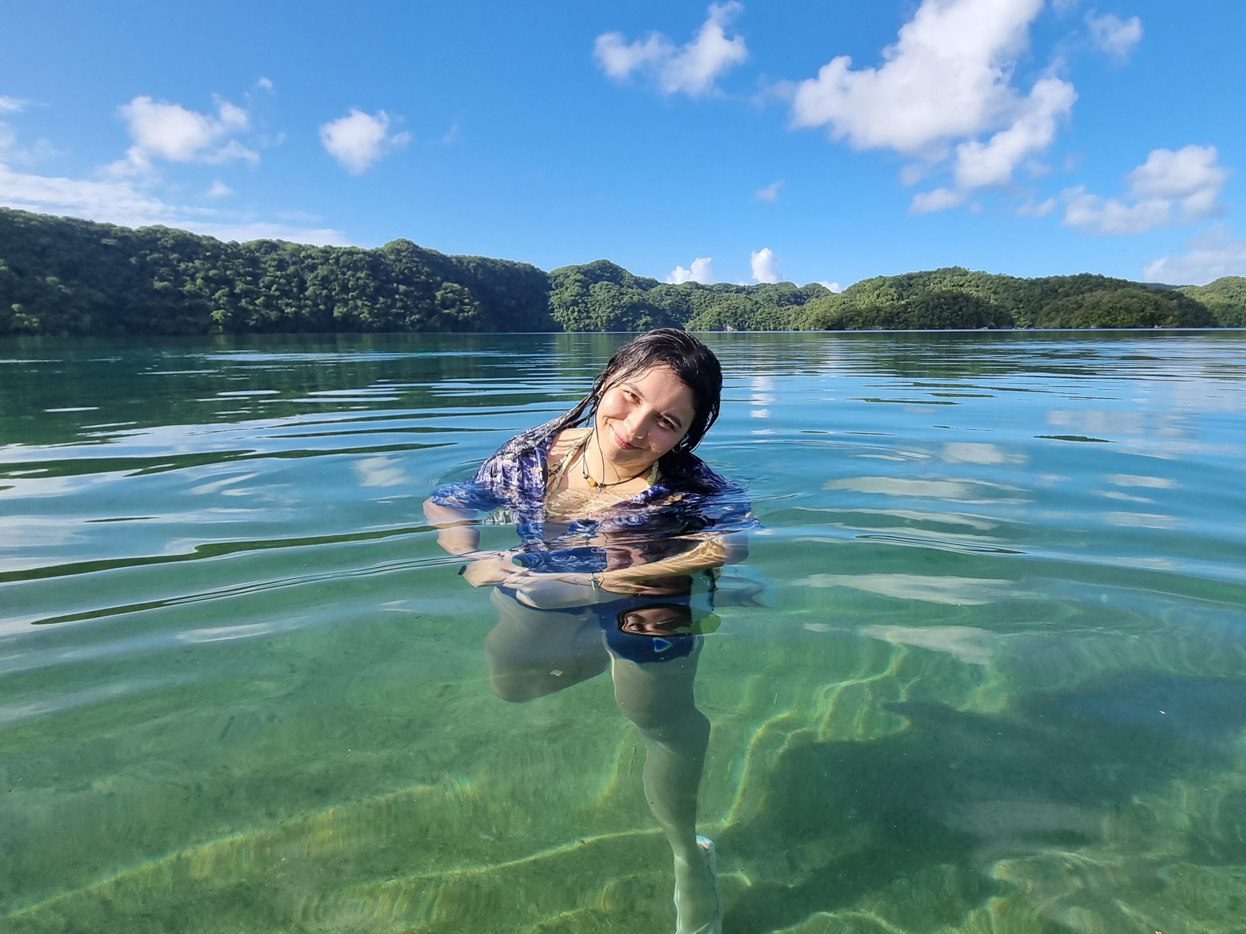 A young woman with wet dark hair smiling and leaning her head to the side while swimming in a clear lake with green hills and a blue sky with white clouds in the background.