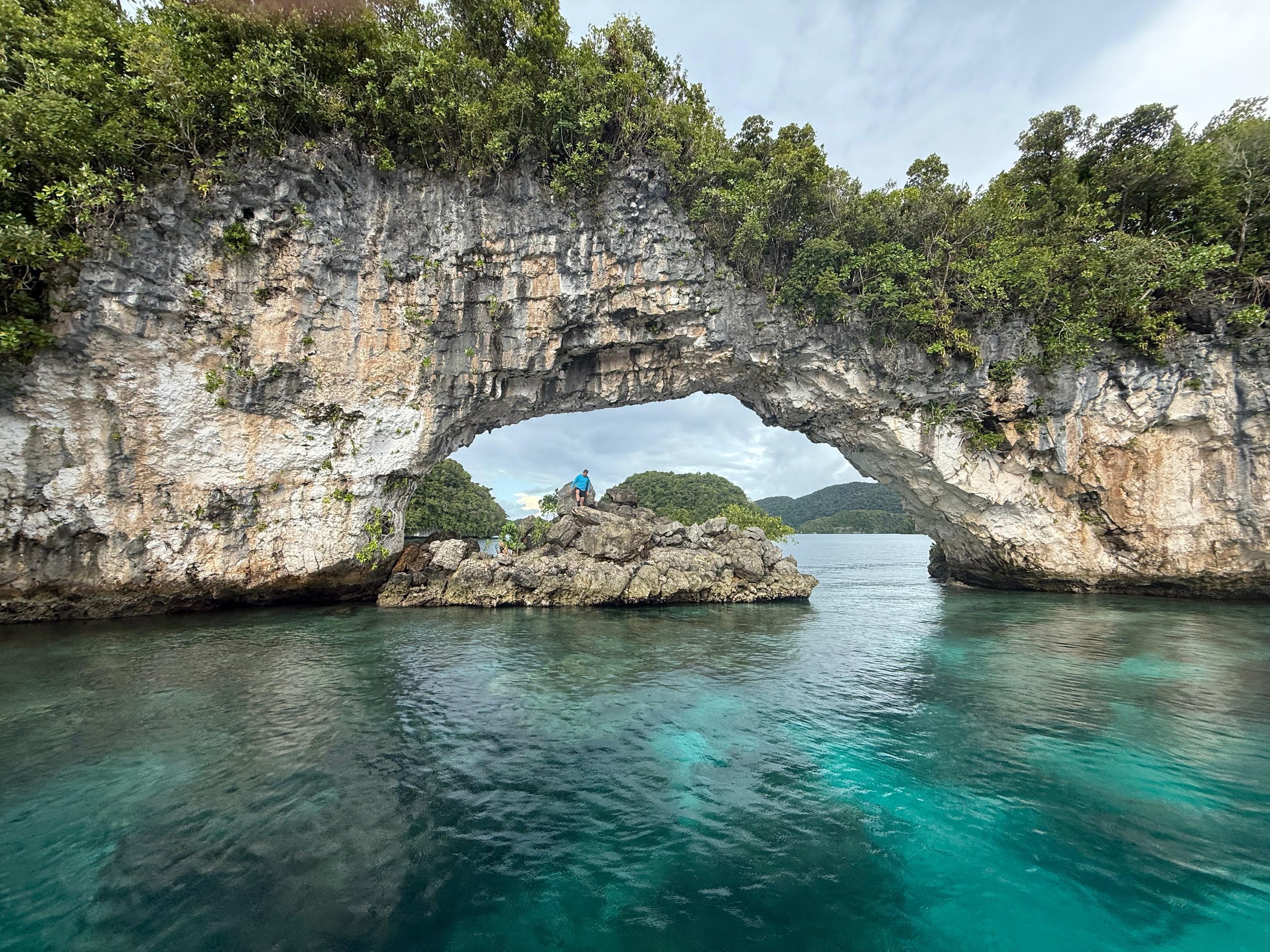 A person hiking on rocks under a large rock arch over turquoise water with green trees and hills in the background.