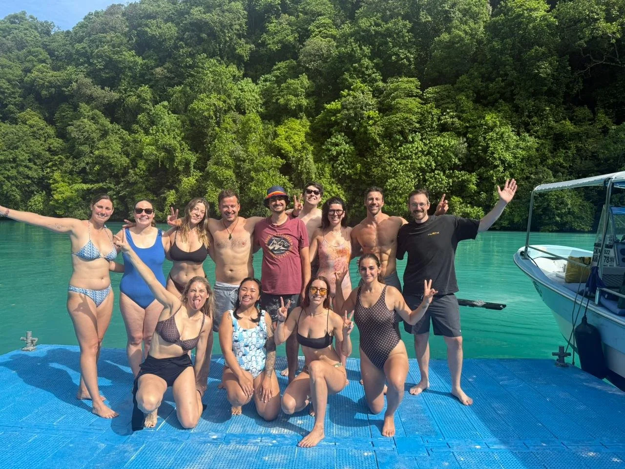 Group of people smiling and posing on a dock near a body of water with a lush green forest in the background. Some are making peace signs, and they are dressed in swimsuits, indicating a swimming or boating outing.