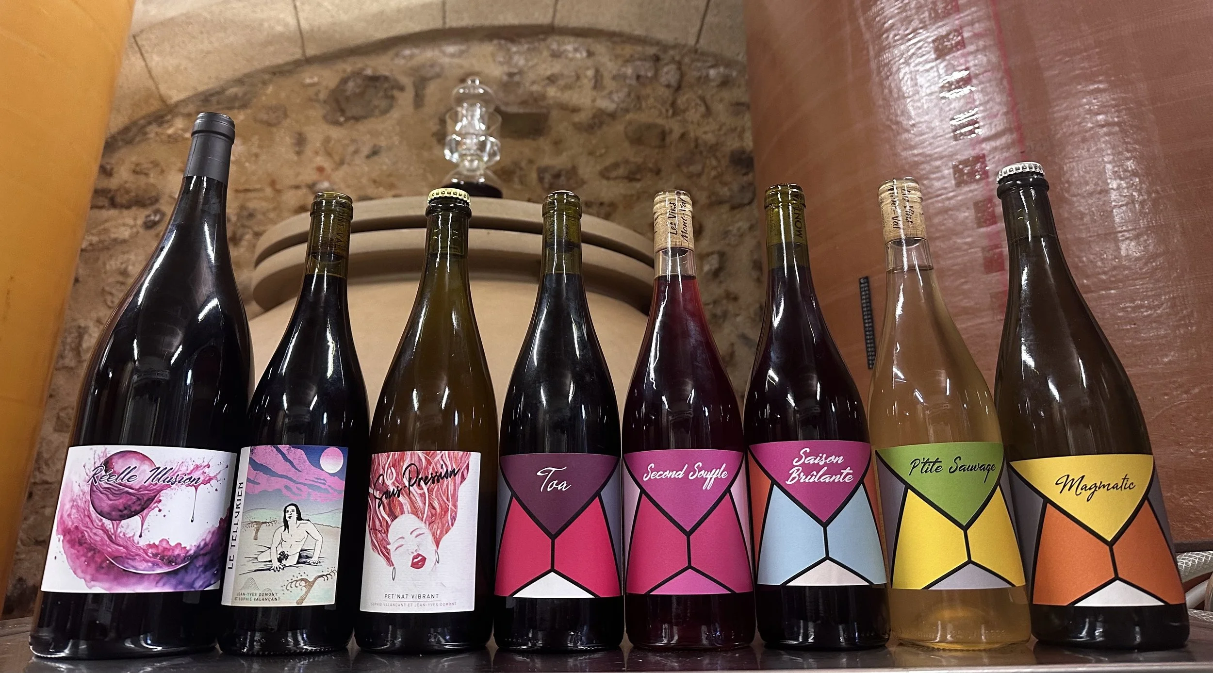 Une rangée de huit bouteilles de vin avec des étiquettes colorées et artistiques, alignées sur une surface en bois, avec un décor de mur en pierre en arrière-plan.