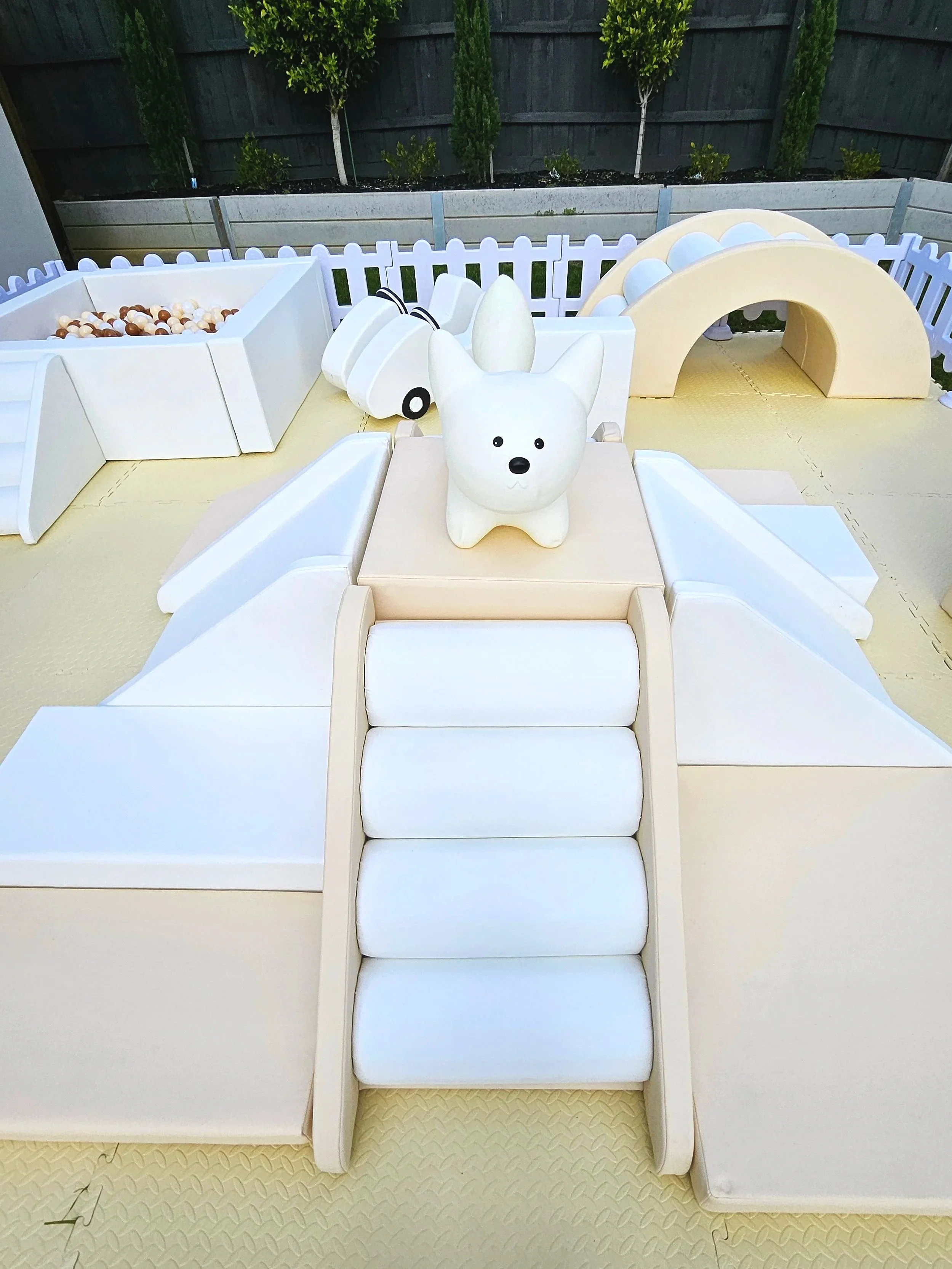 A children's outdoor play area with a dog-shaped speaker, a small slide, a ball pit with brown and white balls, a bridge, and a chain tunnel, all enclosed by a white picket fence, with a garden and black fence in the background.