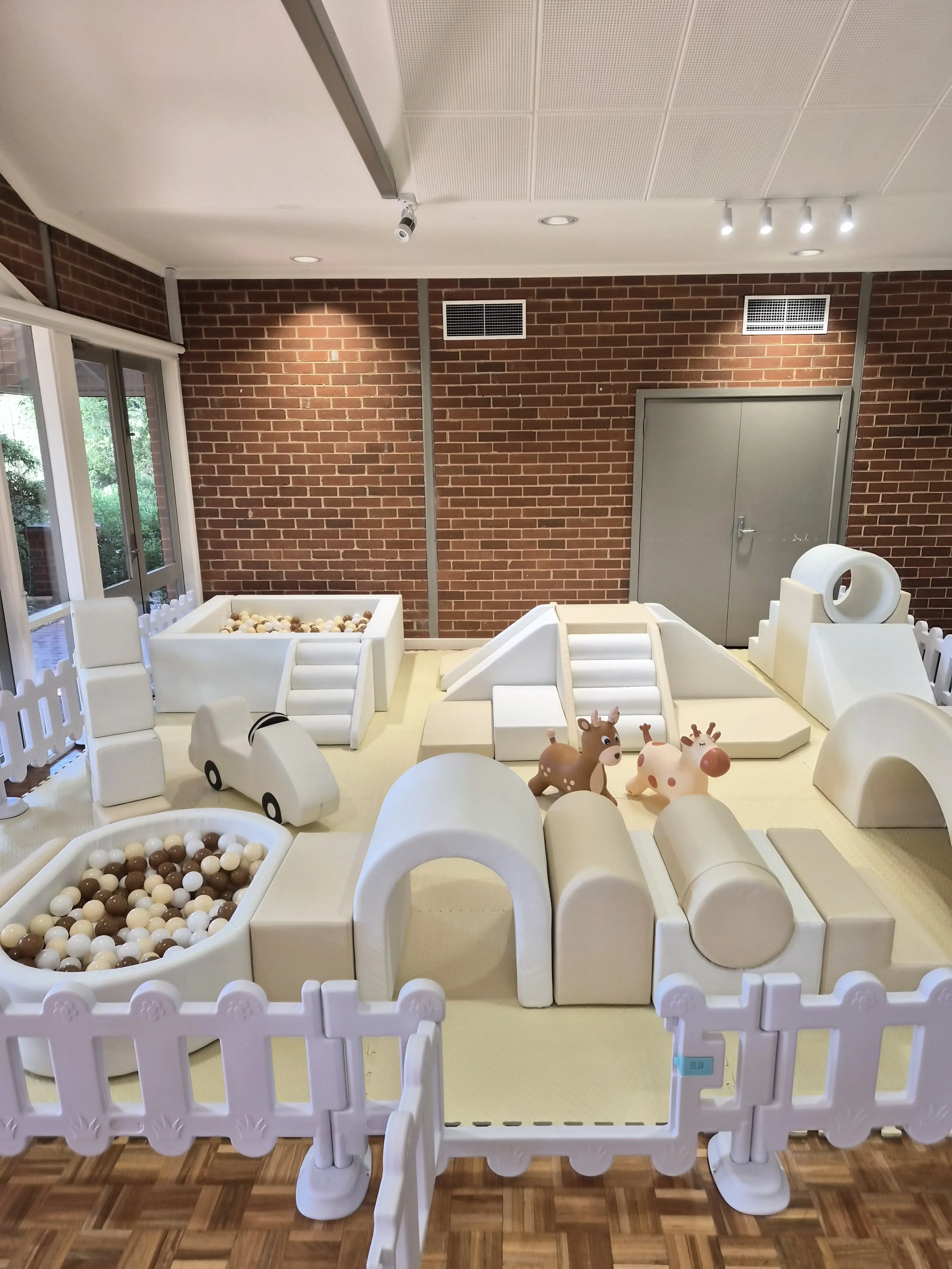 Indoor play area with white plastic slides, ball pits, and animal toys, enclosed by white picket fences, against a brick wall with vents and a gray door.