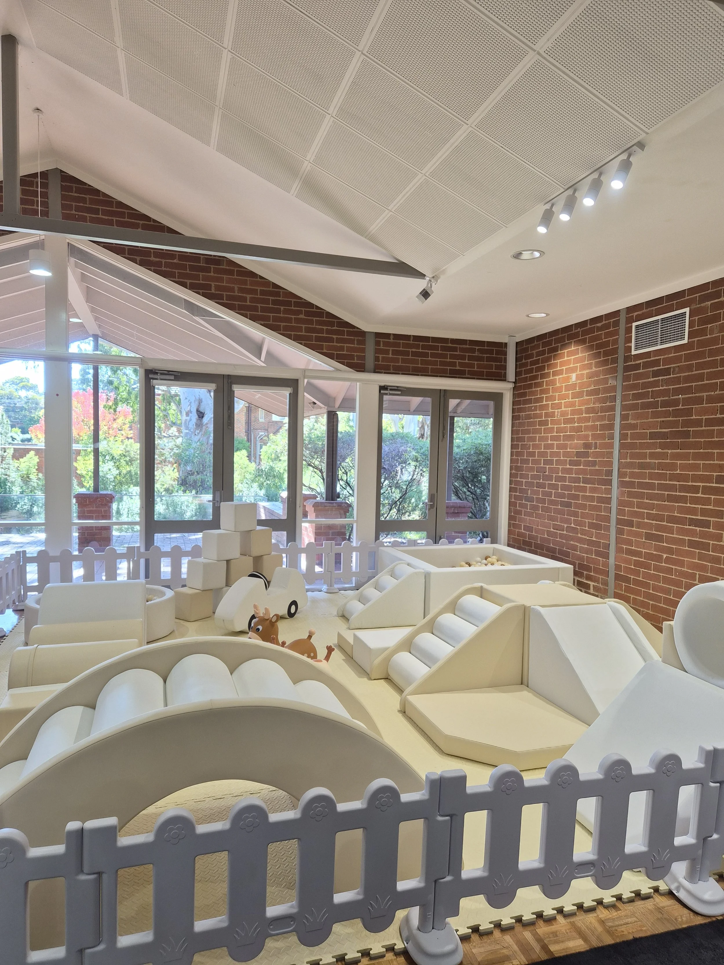 Indoor children’s play area with soft padded flooring, white cave-like structures, a small fence with flower patterns, and toy animals. Large windows in the background show trees and buildings outside.